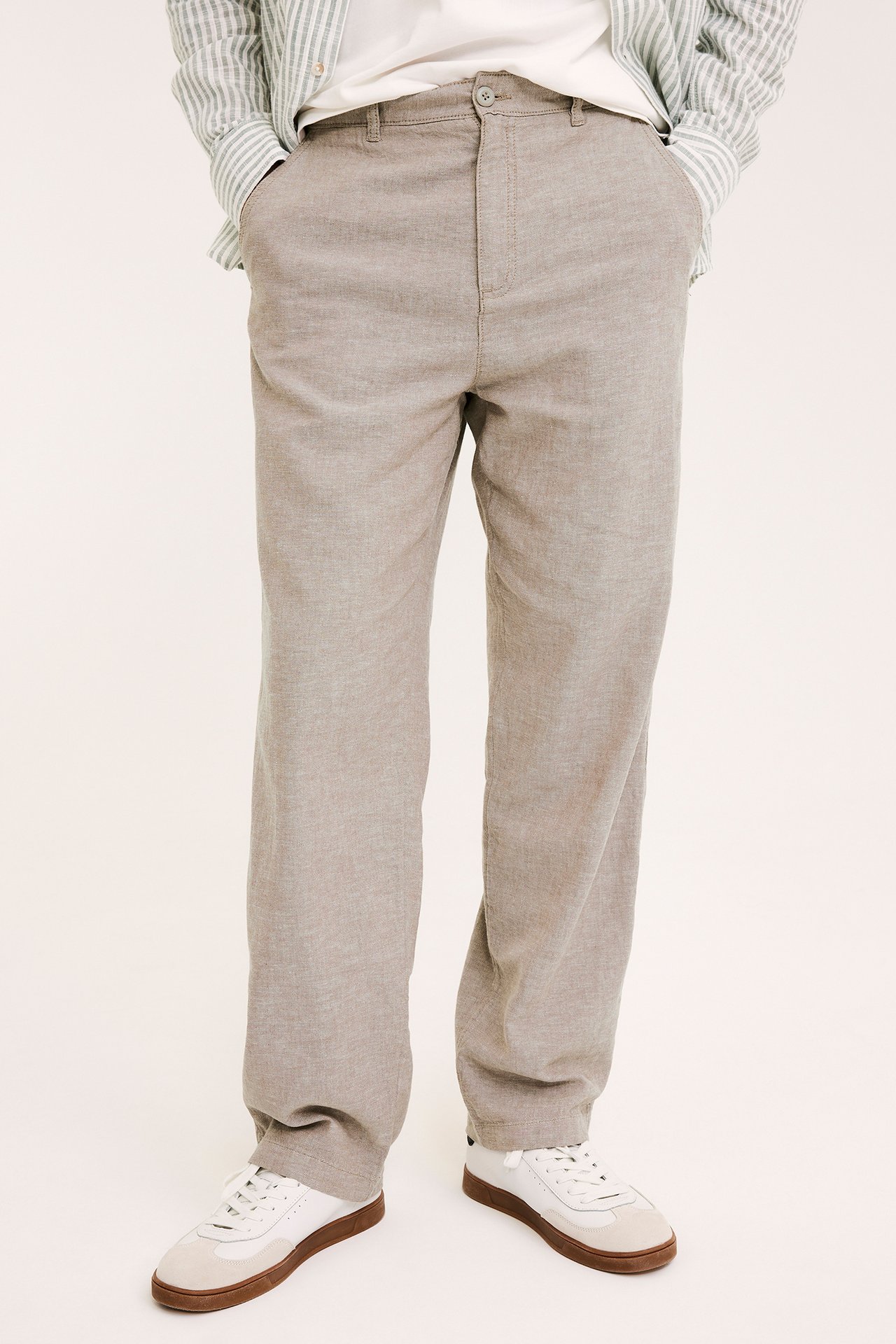 Widok z przodu: mężczyzna w jasnobrązowych spodniach lnianych, białym t-shircie, koszuli w paski i białych tenisówkach.