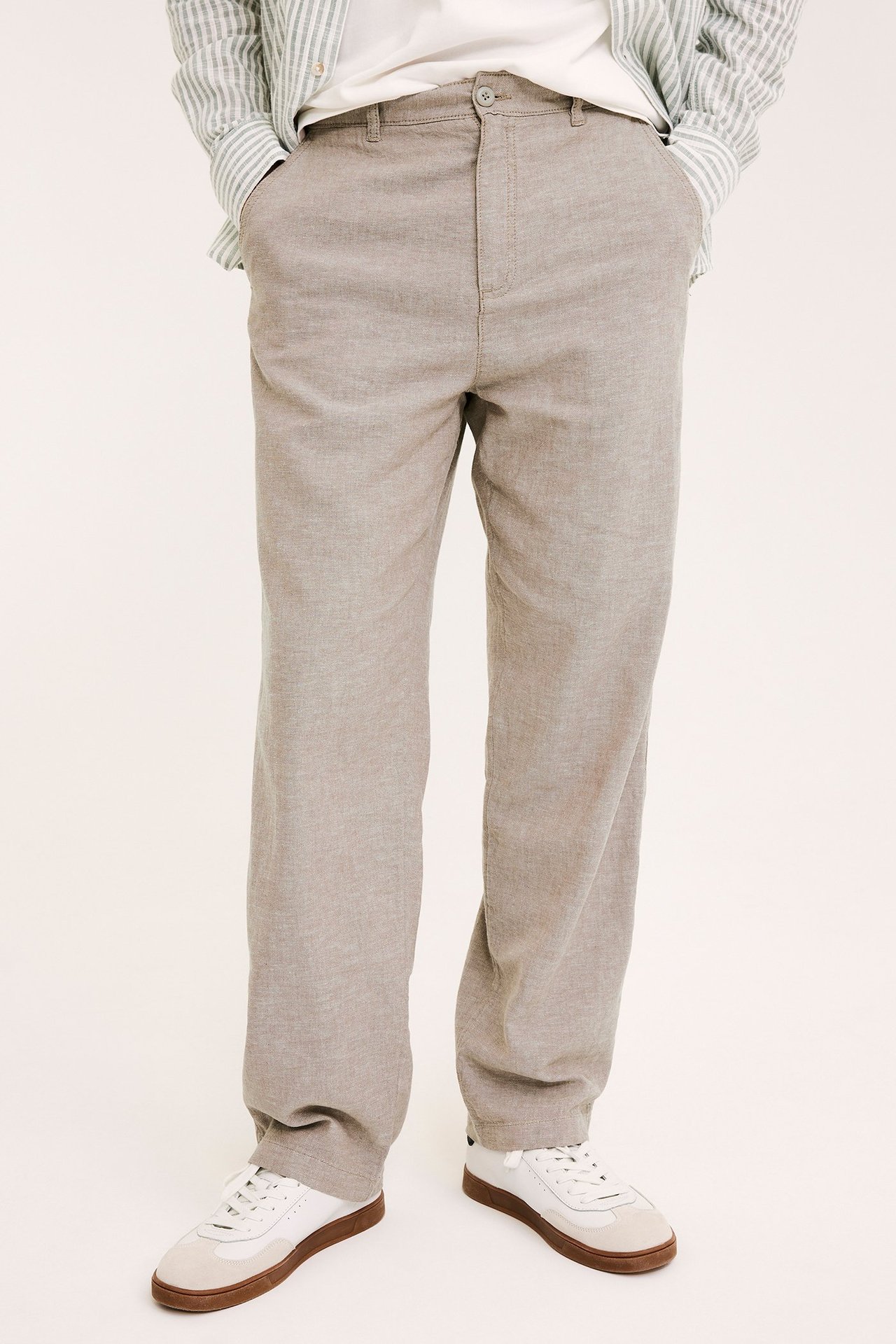 Miehen etunäkymä: yllään vaaleanruskeat pellavasekoite housut, valkoinen t-paita, raidallinen paita ja valkoiset lenkkarit.