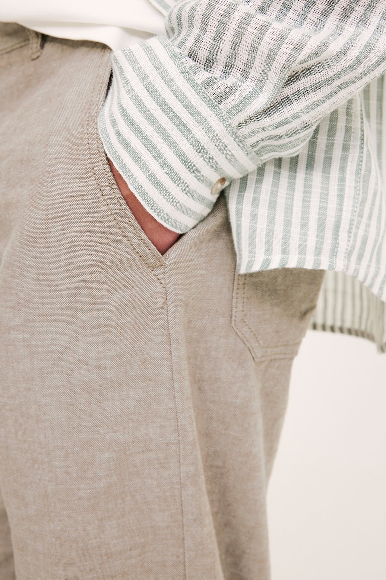 Zbliżenie mężczyzny w jasnobrązowych spodniach z mieszanki lnu, ręka w kieszeni, w koszuli w paski.
