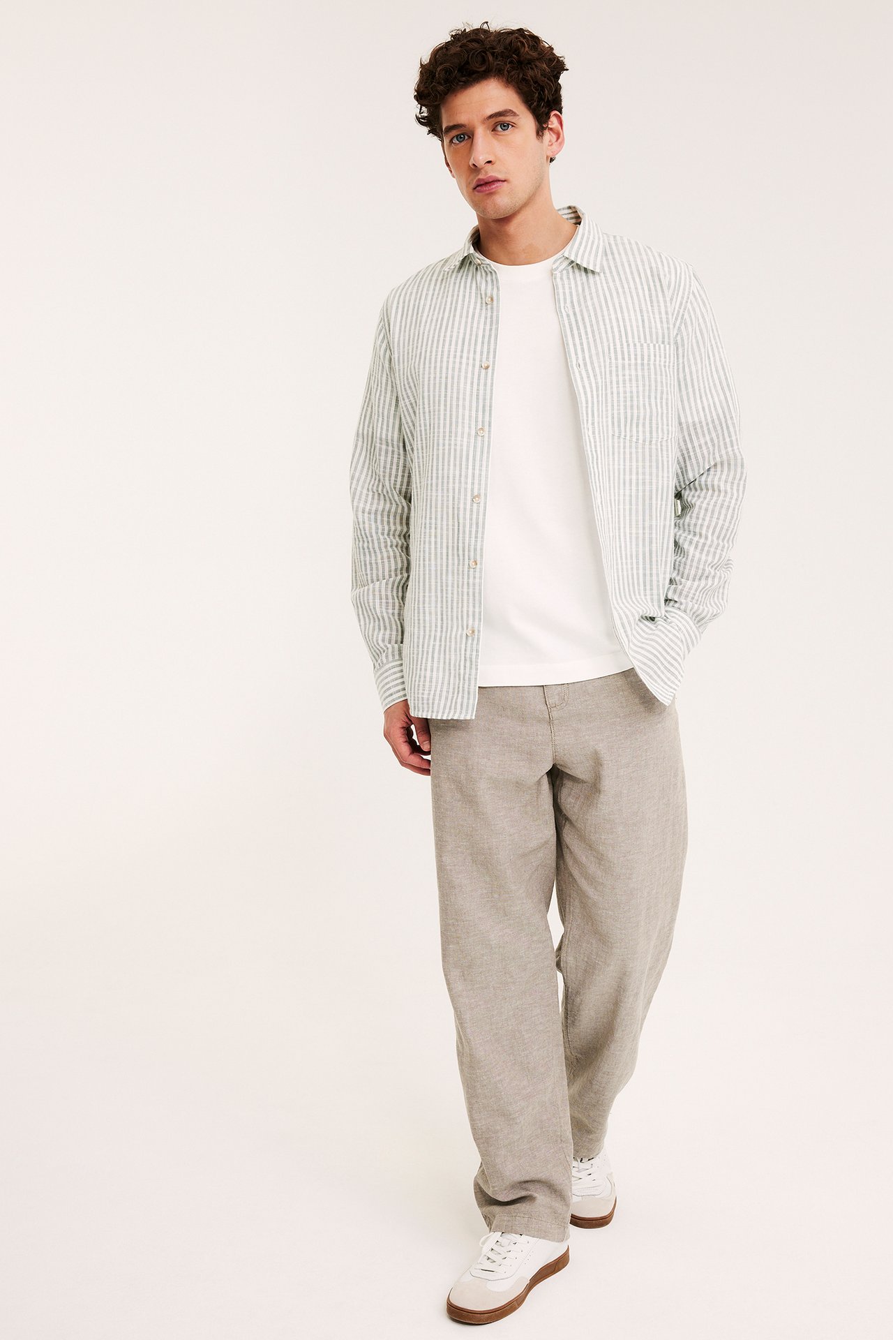 Widok z przodu: Mężczyzna w jasnobrązowych lnianych spodniach regular fit, białym t-shircie, rozpiętej koszuli w paski i białych sneakersach.