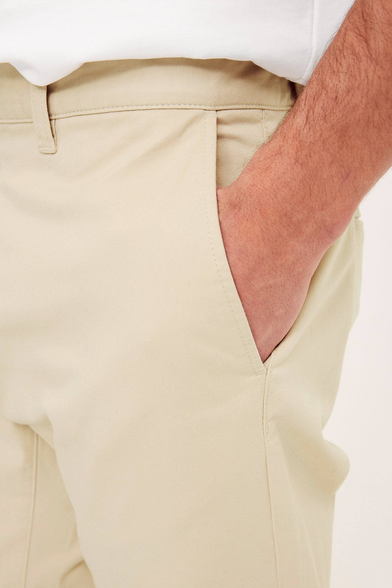 Nærbilde forfra: beige regular fit bomullstwill chinos, manns hånd i lommen, over hvit t-skjorte.