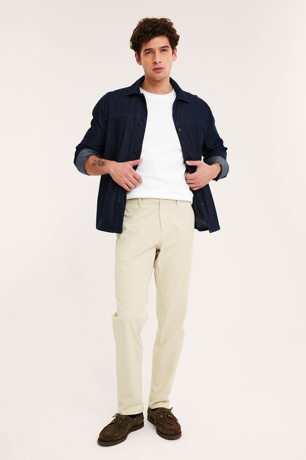 Framifrån: Man i beige regular fit chinos, vit t-shirt, mörkblå skjorta, bruna seglarskor.