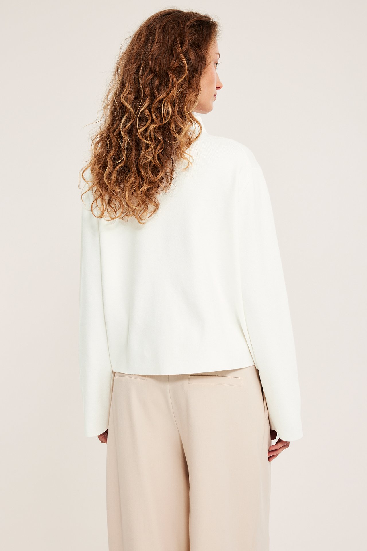 Widok z tyłu: kobieta w kremowym swetrze z kołnierzem i luźnych beżowych spodniach.