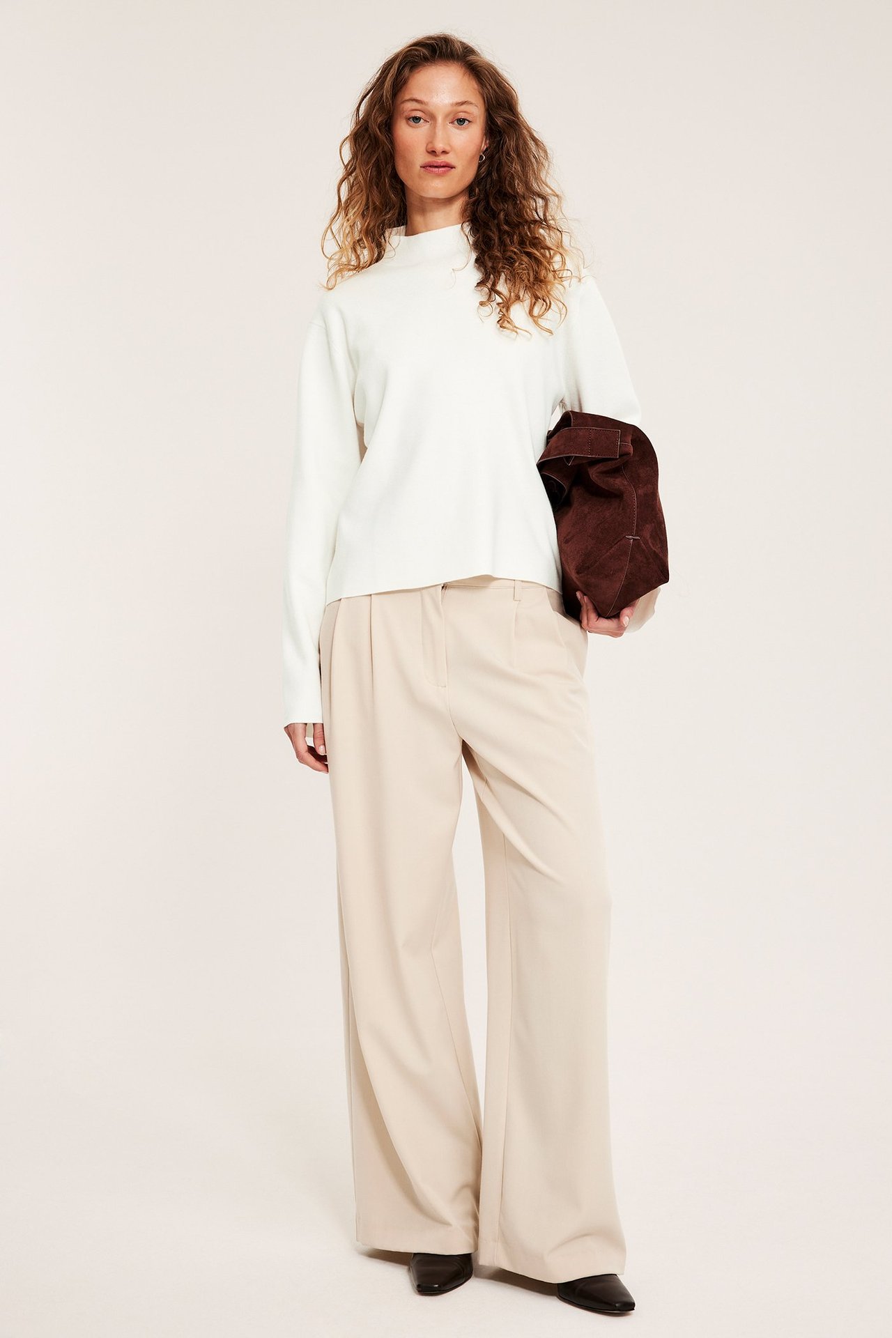 Edestäpäin: nainen luonnonvalkoisessa kaulusneuleessa, vaaleanbeigeissä housuissa, ruskea mokkalaukku kädessä.