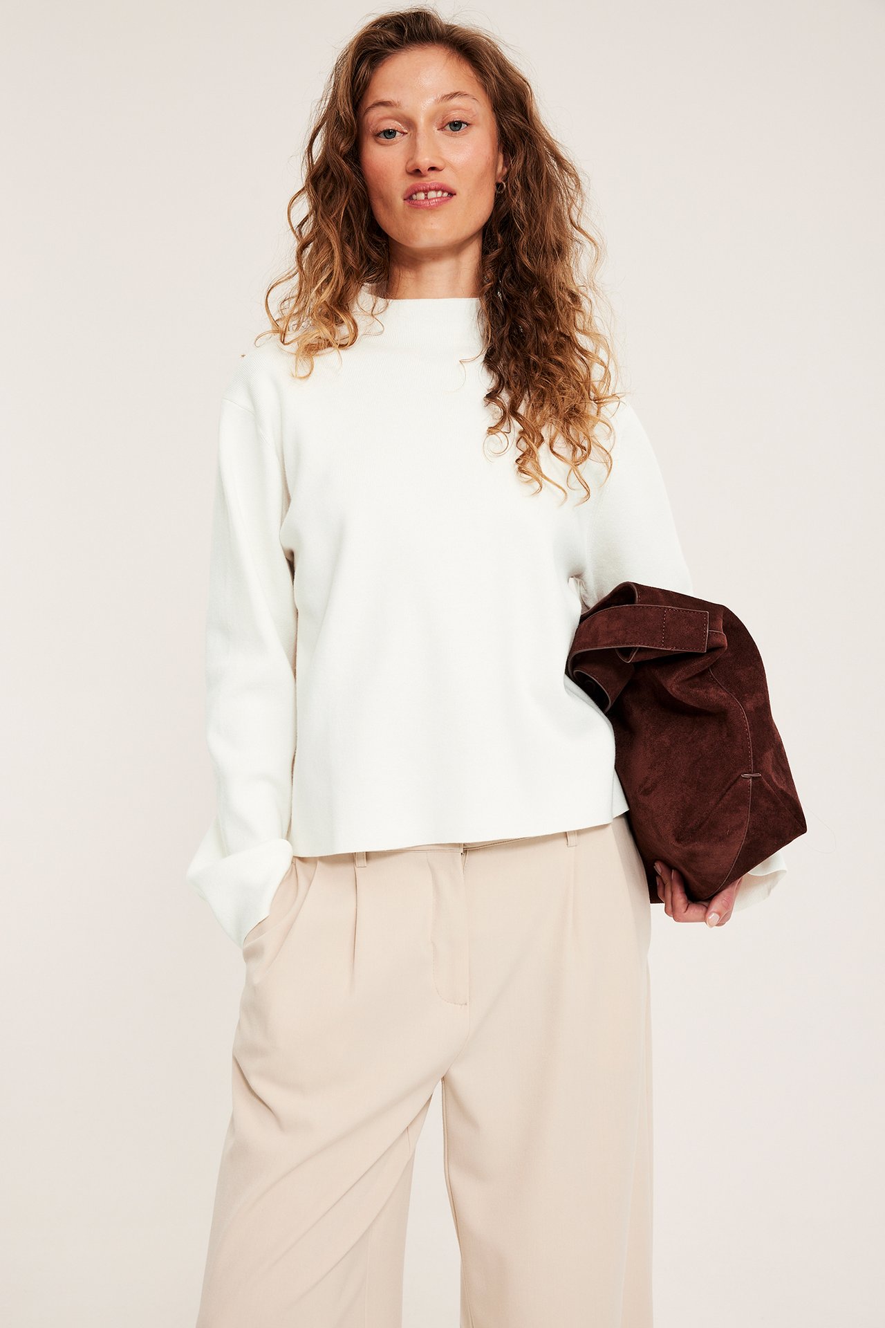 Widok z przodu: kobieta w łamanobiałym swetrze z kołnierzem, beżowych spodniach, trzymająca brązową torbę.