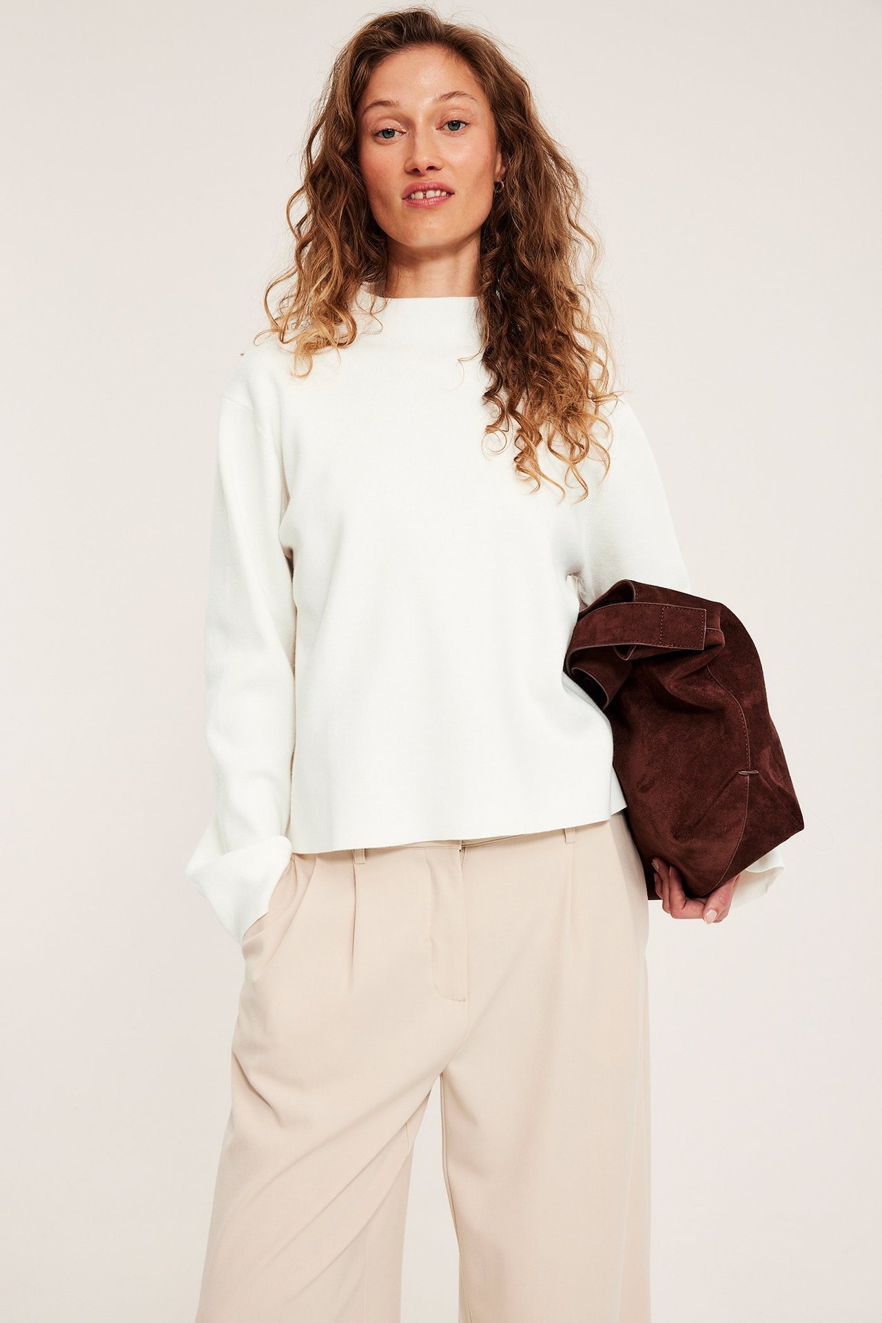 Widok z przodu: kobieta w łamanobiałym swetrze z kołnierzem, beżowych spodniach, trzymająca brązową torbę.
