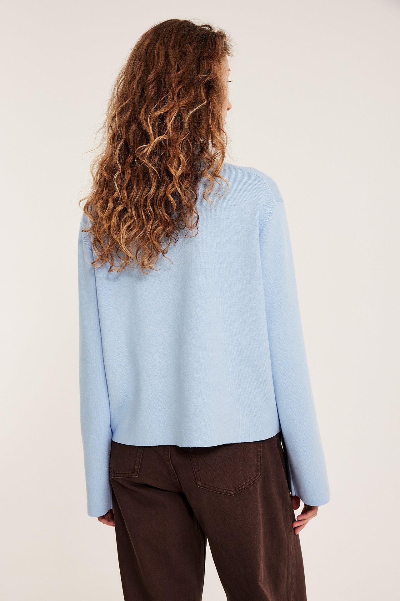 Tyłem: Kobieta w jasnoniebieskim swetrze z kołnierzem i ciemnobrązowych szerokich spodniach.