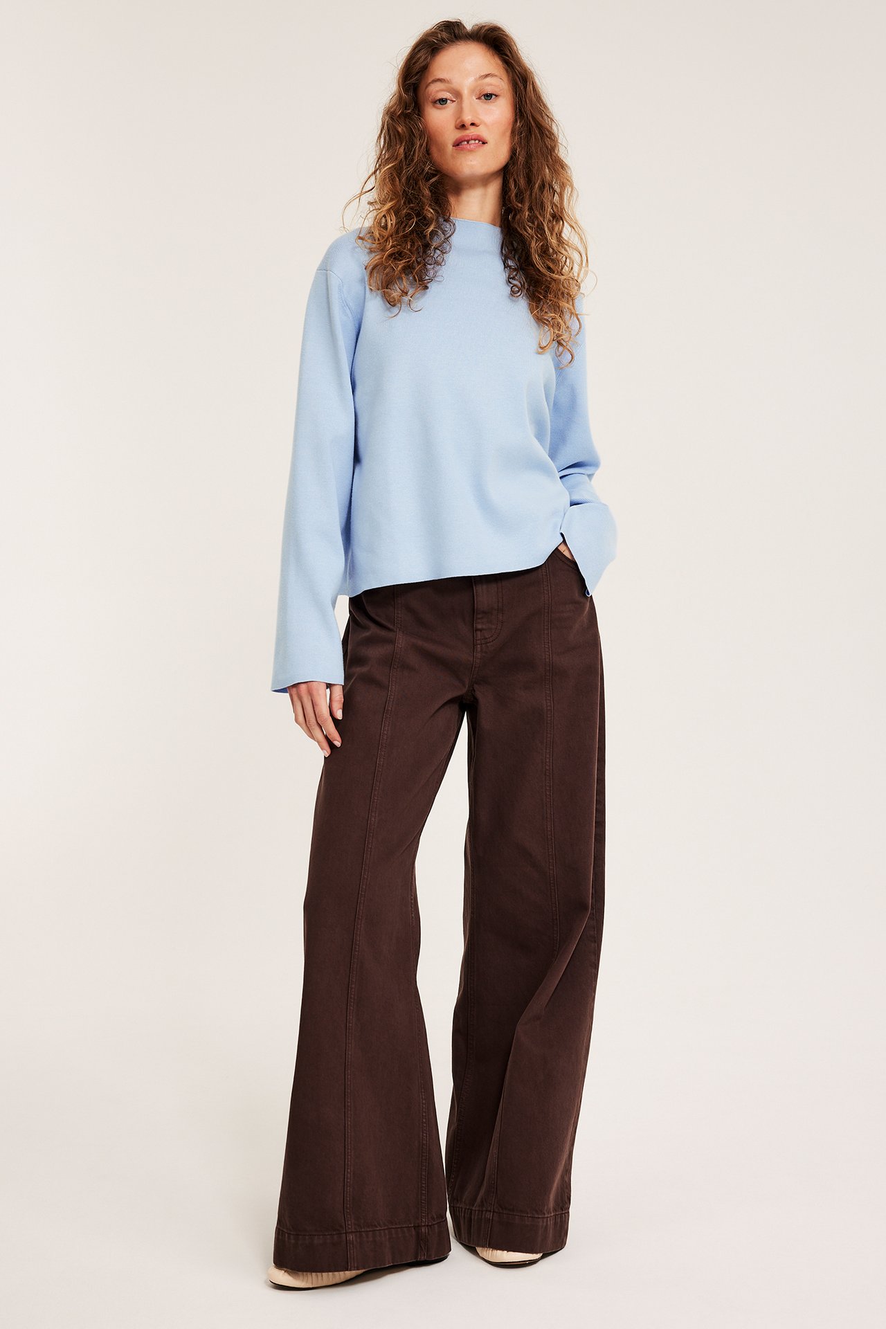 Widok z przodu: kobieta w jasnoniebieskim swetrze z golfem i ciemnobrązowych szerokich spodniach.