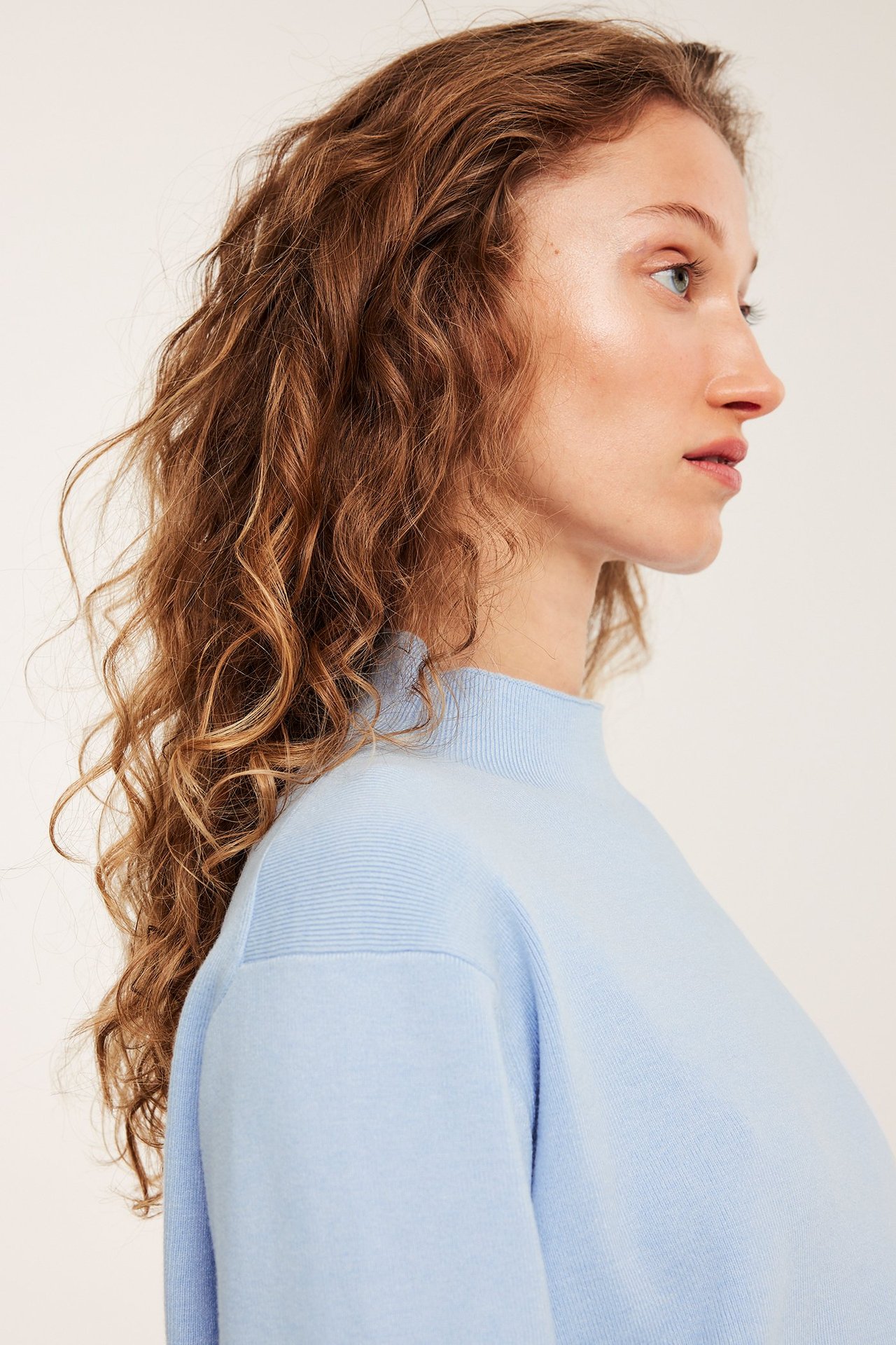 Kobieta z boku w jasnoniebieskim swetrze z dzianiny, z wysokim kołnierzem i prążkowanymi ramionami.