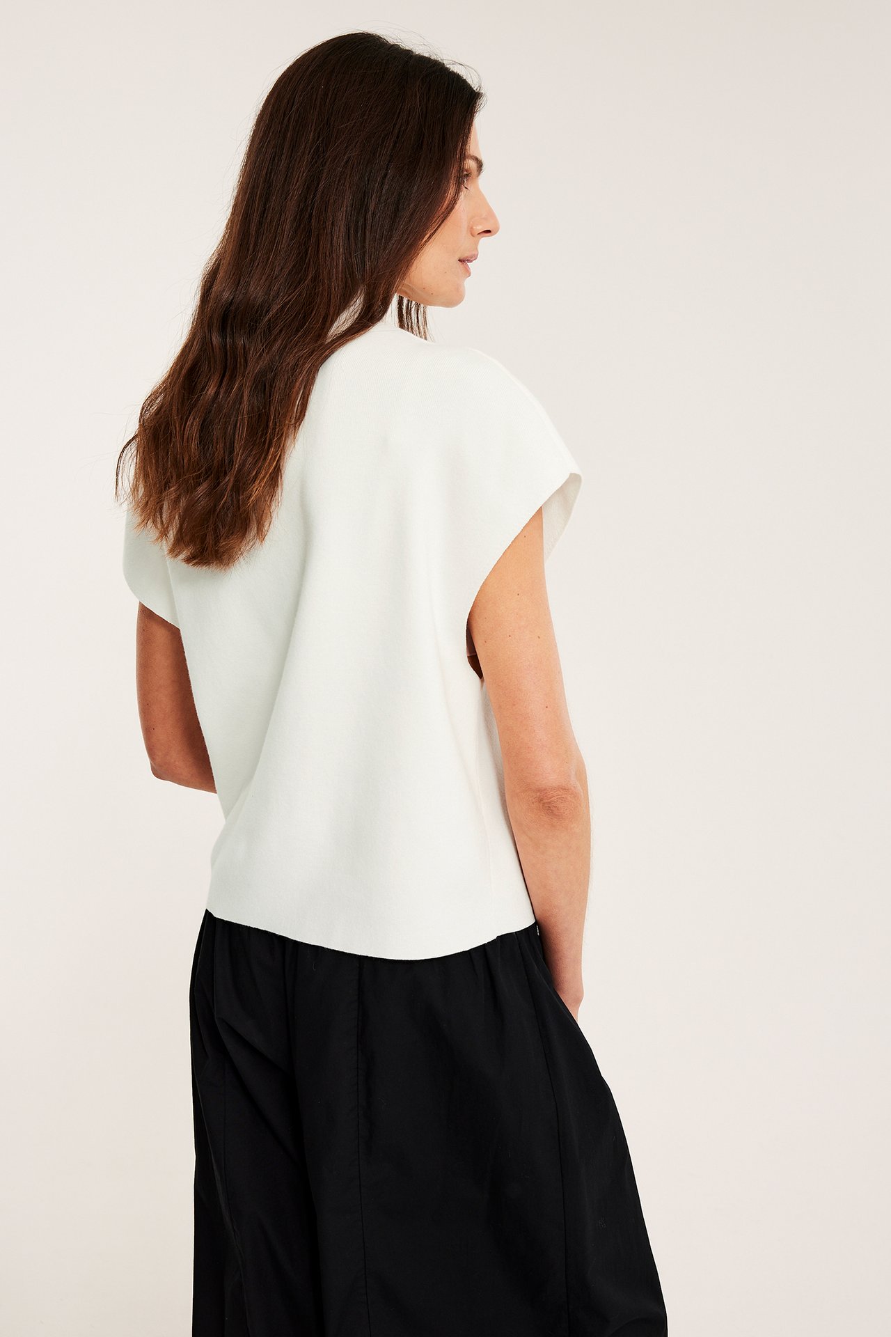 Widok z boku kobiety w kremowej dzianinowej kamizelce ze stójką i szerokich czarnych spodniach.