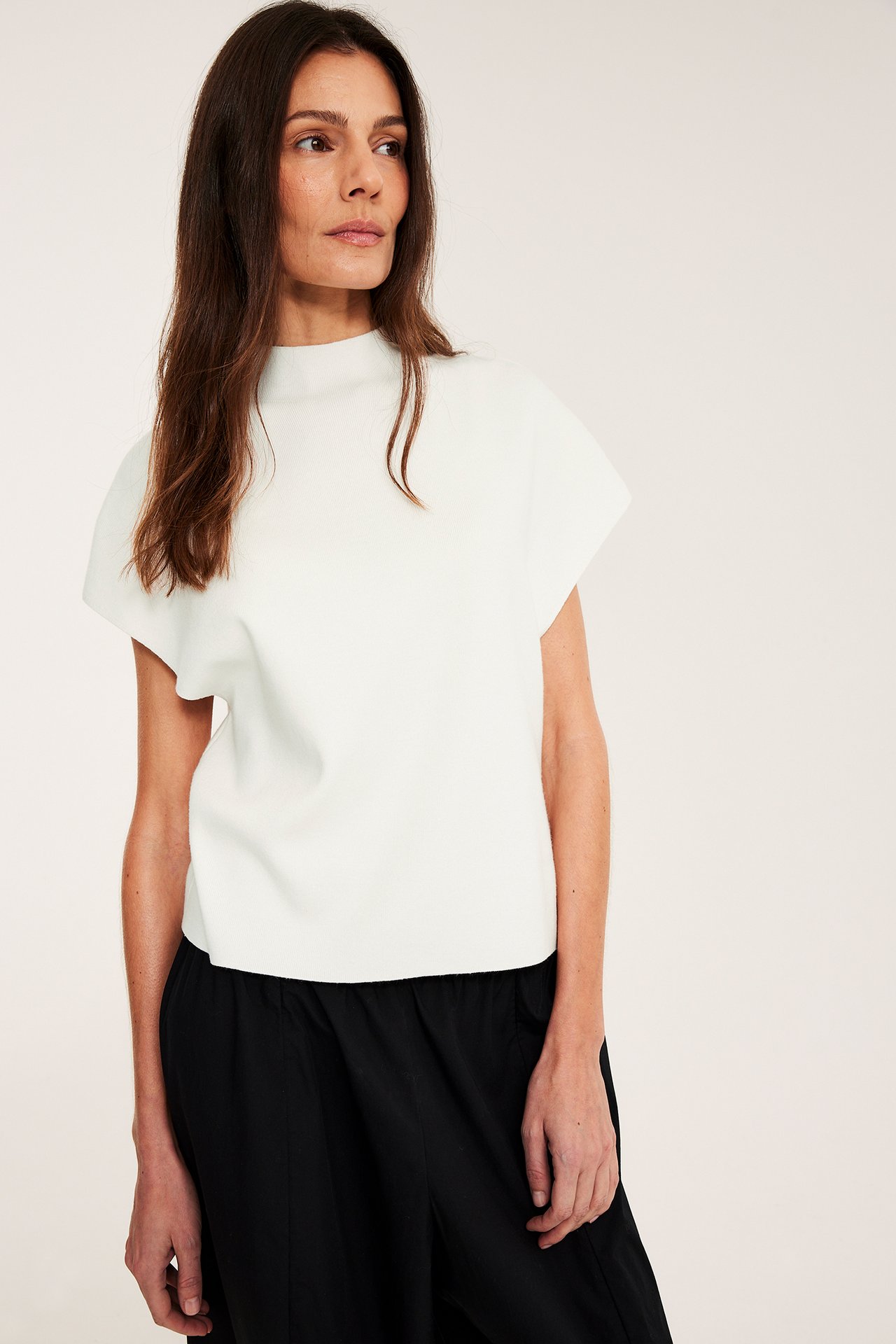 Naisen etunäkymä, yllään luonnonvalkoinen neulottu liivi, jossa on kaulus ja mustat leveälahkeiset housut.