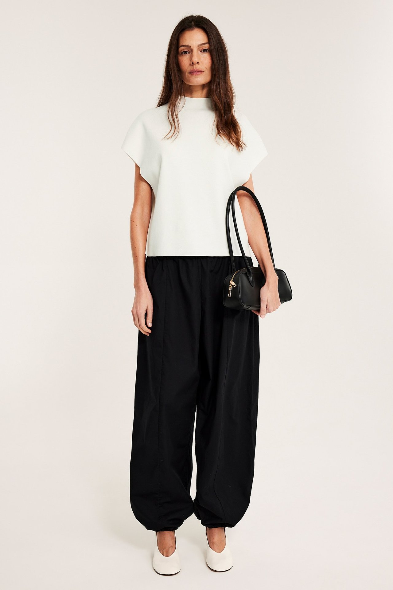 Kobieta w beżowym dzianinowym topie z kołnierzem i krótkimi rękawami, czarnych szerokich spodniach, białych butach, trzymająca czarną torebkę.