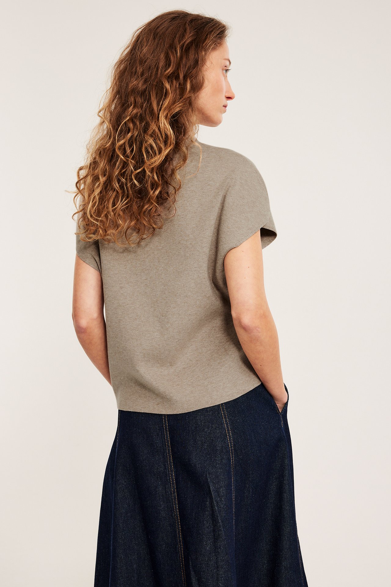 Sivukuva: nainen beigenvärisessä meleeratussa lyhythihaisessa neuleliivissä, jossa kaulus, ja tummansinisessä farkkuhameessa.