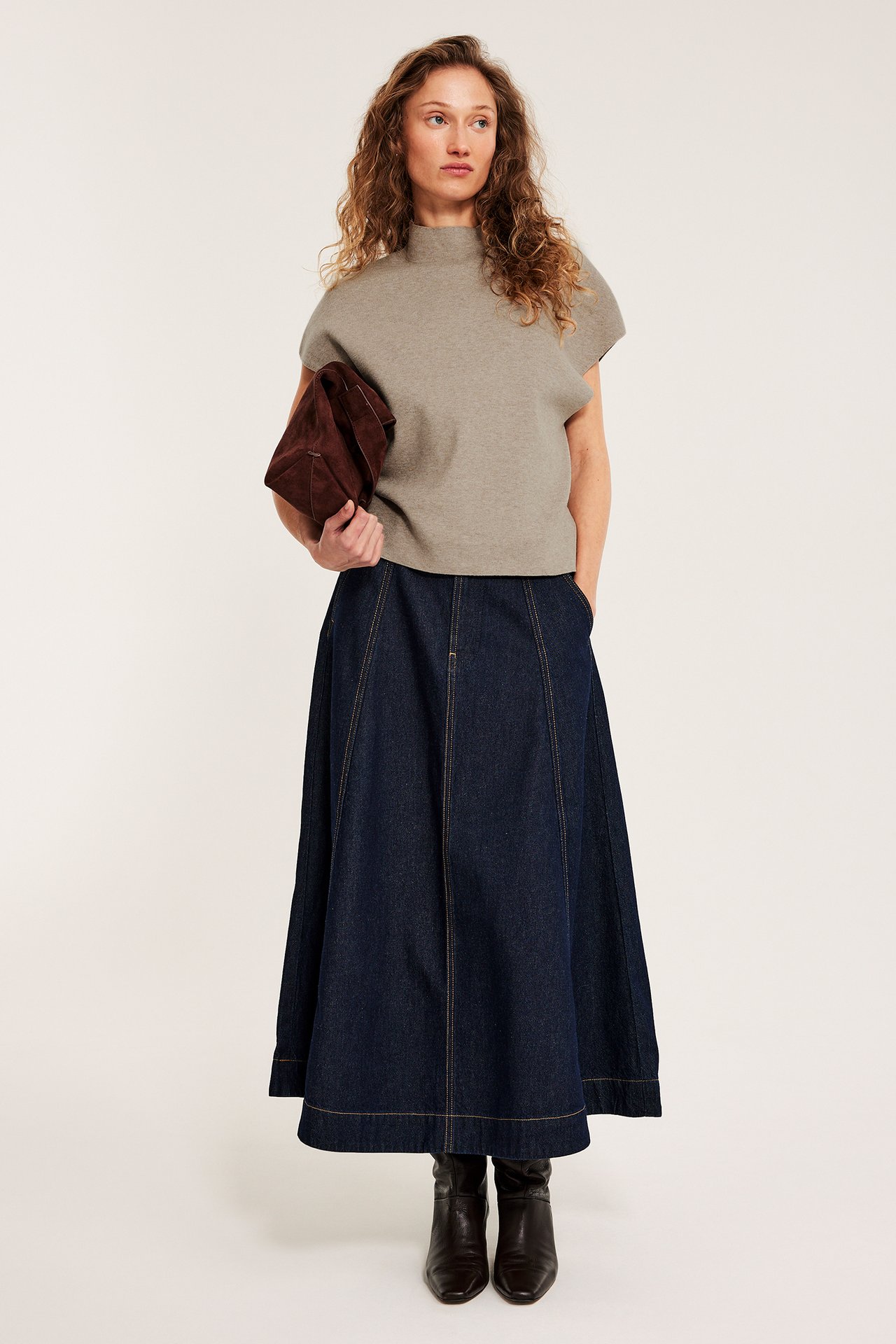 Nainen melange beigessä kauluksellisessa neuleliivissä, tummansinisessä farkkuhameessa, ruskeissa saappaissa, pitäen ruskeaa laukkua.