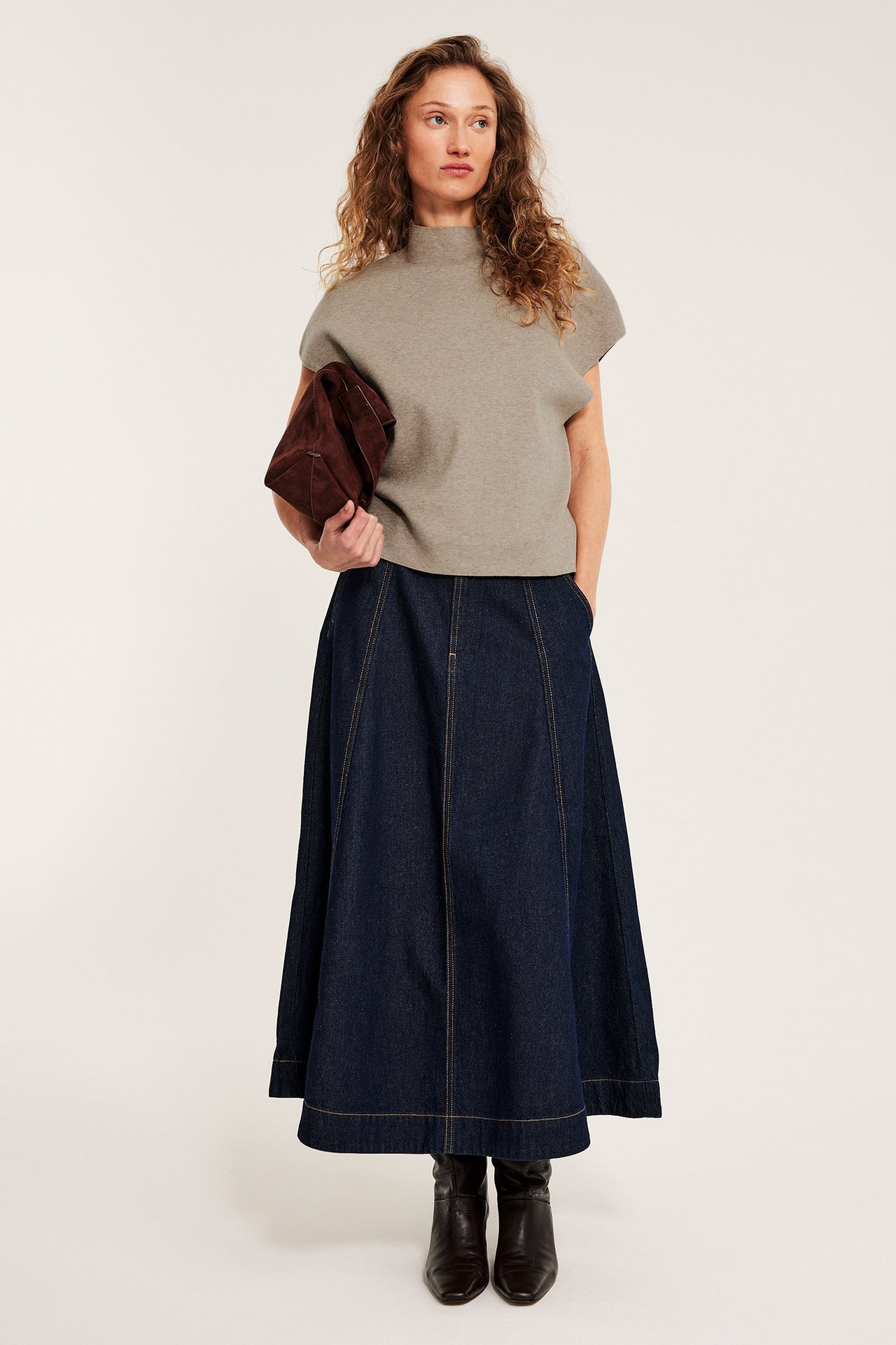 Kvinne i melert beige strikket vest med krage, mørkeblått denimskjørt, brune støvler, holder brun veske.