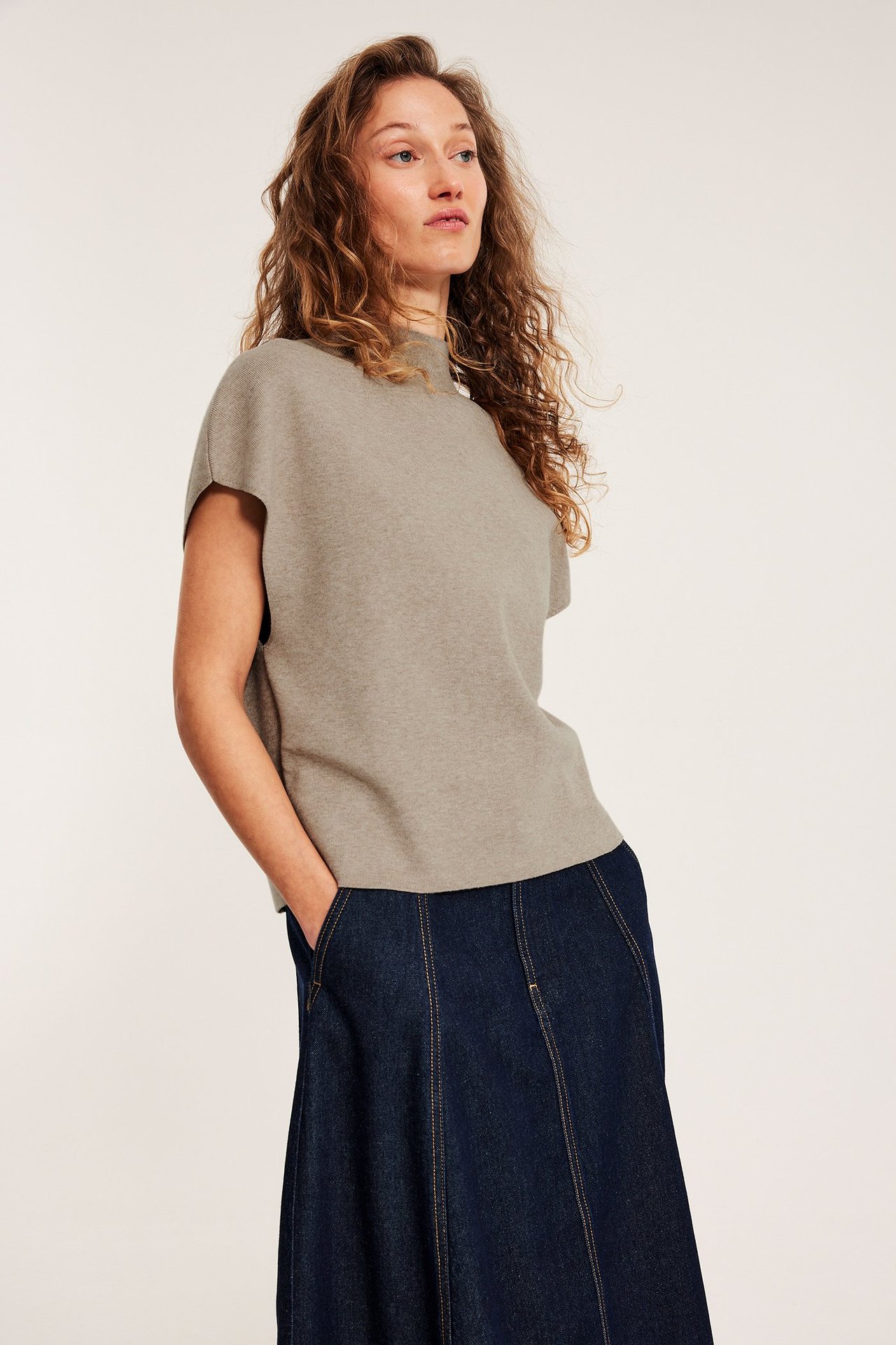 Frontbilde: kvinne i melert beige strikket vest med høy krage over mørkeblått denim A-linjeskjørt.