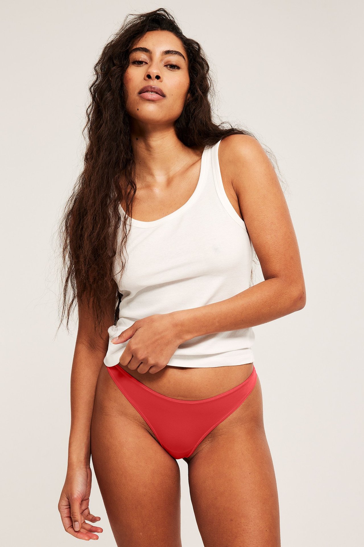 Forfra, en kvinne har på seg røde, ensfargede, supermyke stringtruser og en hvit ribbet singlet.