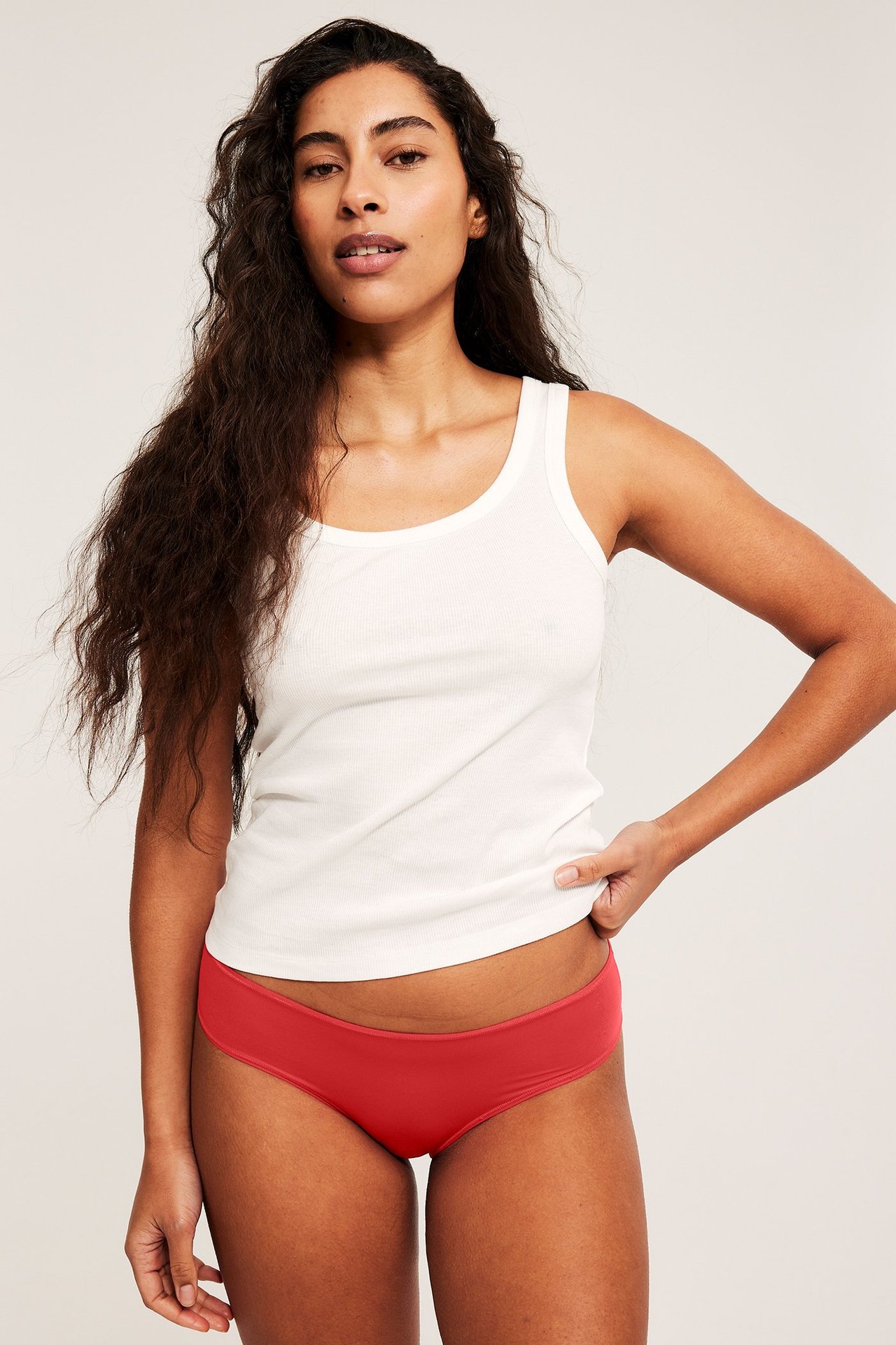 Frontbilde av en kvinne i myke, røde brazilian-truser og en hvit ribbet singlet.