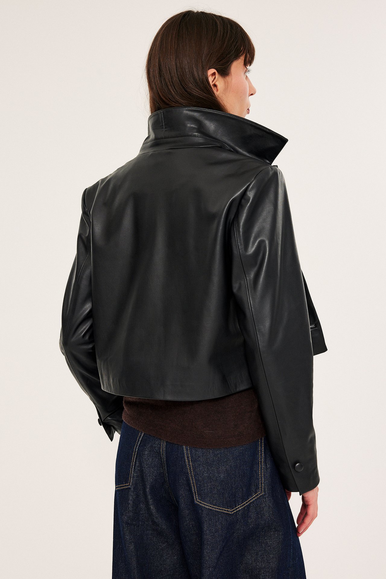 Rygg-sidebilde av kvinne i svart kort jakke i skinnimitasjon over brun topp og mørkeblå jeans.