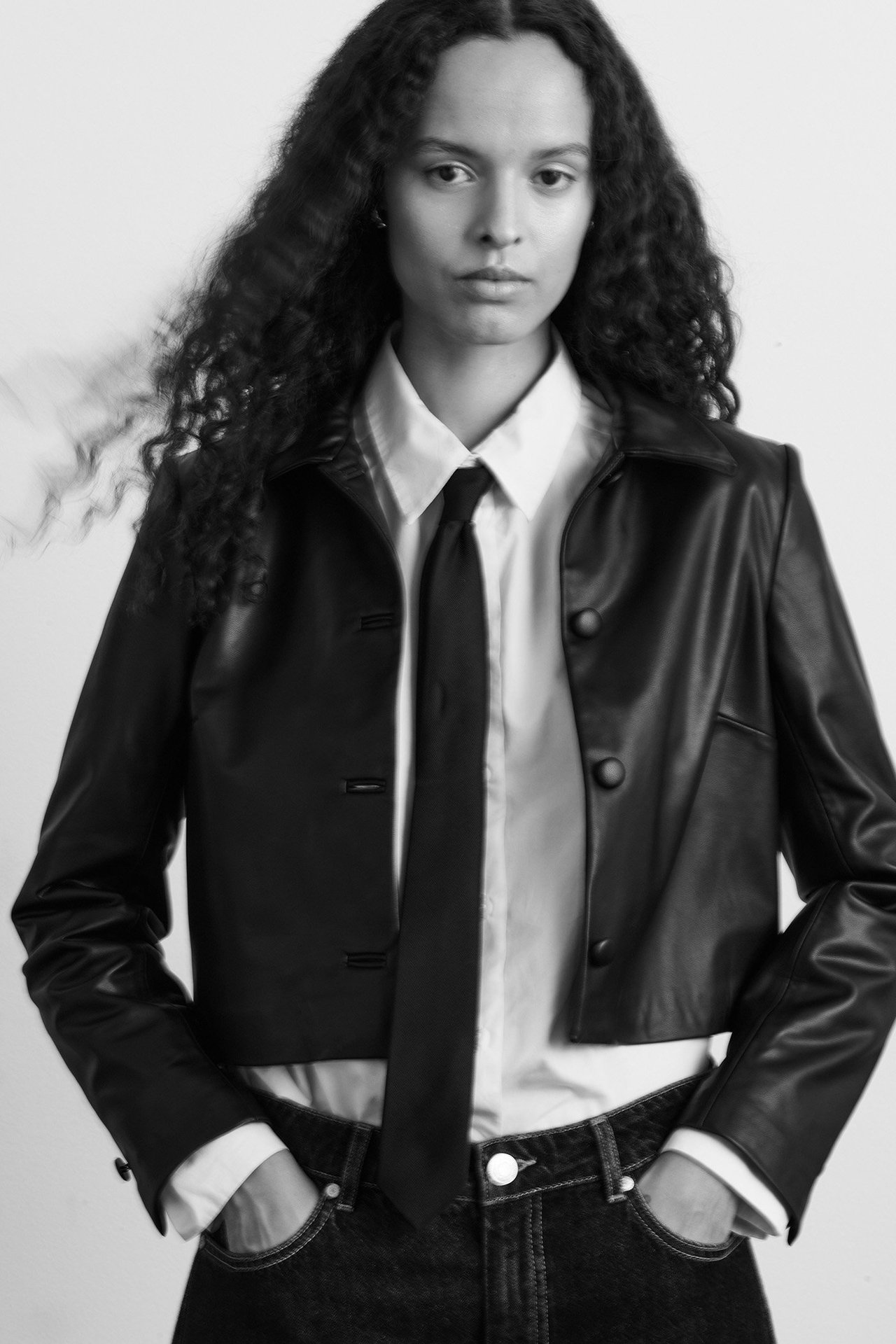 Framifrån: kvinna i svart kort jacka i skinnimitation över vit skjorta, svart slips och mörka jeans.