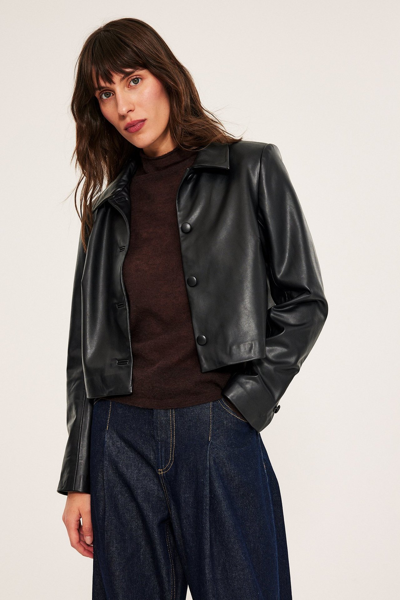 Edestä: nainen mustassa lyhyessä tekonahkatakissa, alla tummanruskea toppi ja leveälahkeiset farkut.