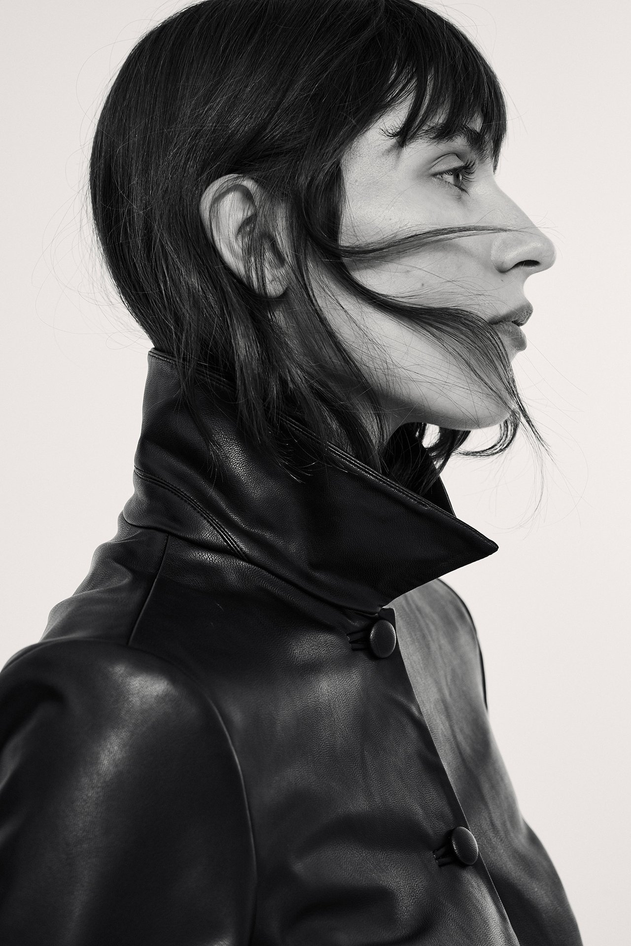 Nærbilde i sideprofil av kvinne i svart kort jakke i imitert skinn med knapper.