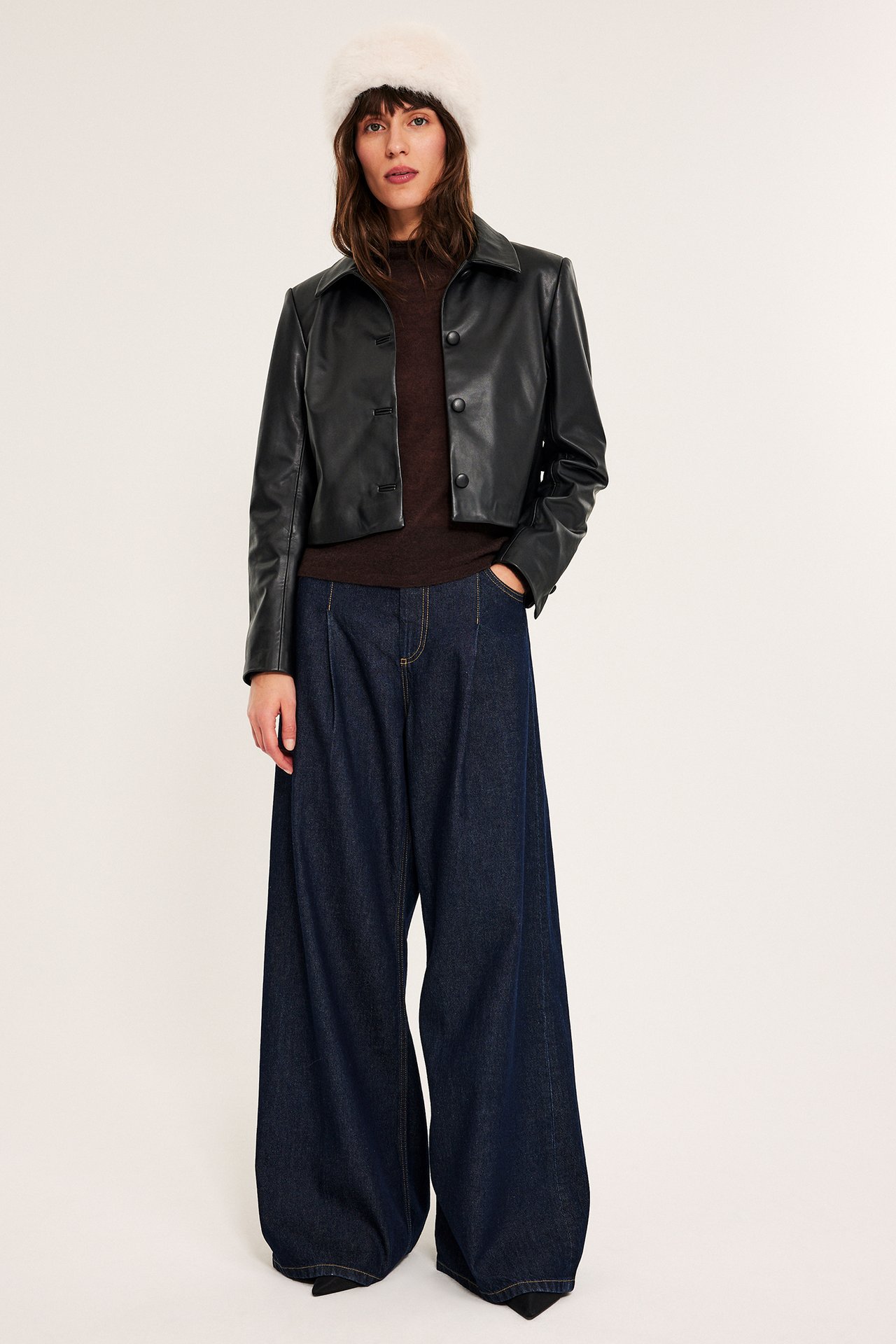 Kvinne har på seg en svart kort jakke i skinnimitasjon, mørkebrun topp, vide blå jeans og en hvit fuskepelslue.