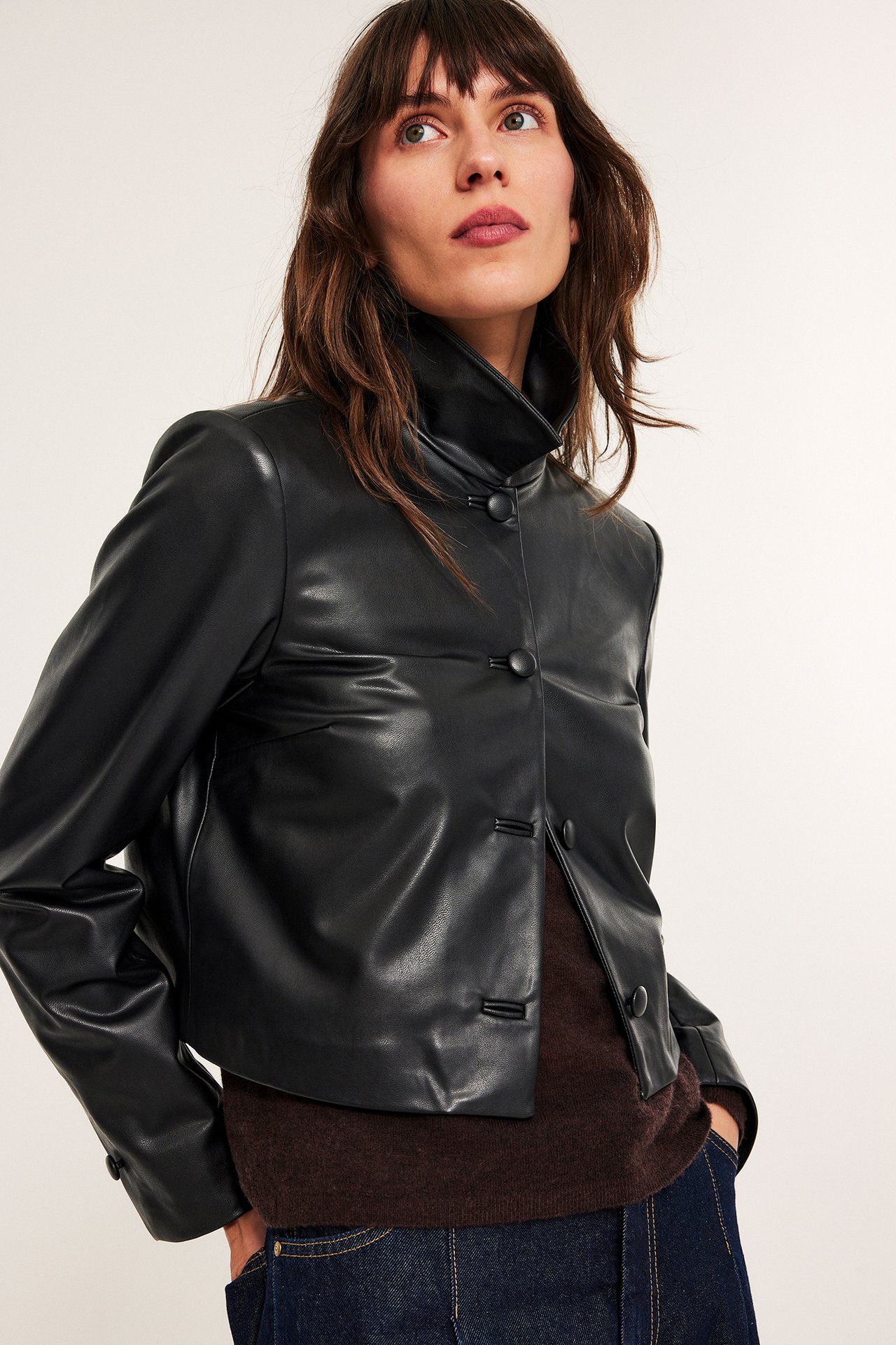 Nainen edestäpäin, musta lyhyt tekonahkatakki napeilla, tummanruskea neule ja tummansiniset farkut.