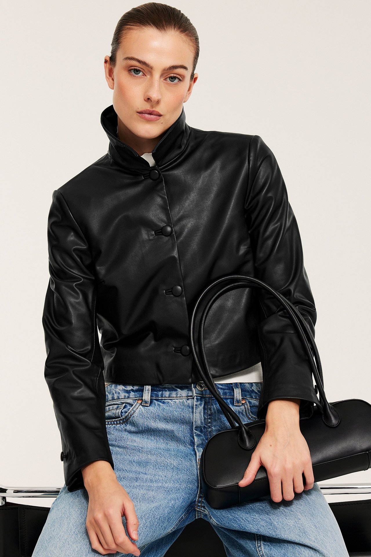 Forfra: kvinne i svart kort PU-jakke med knappelukking, lyseblå jeans, holder en svart håndveske.