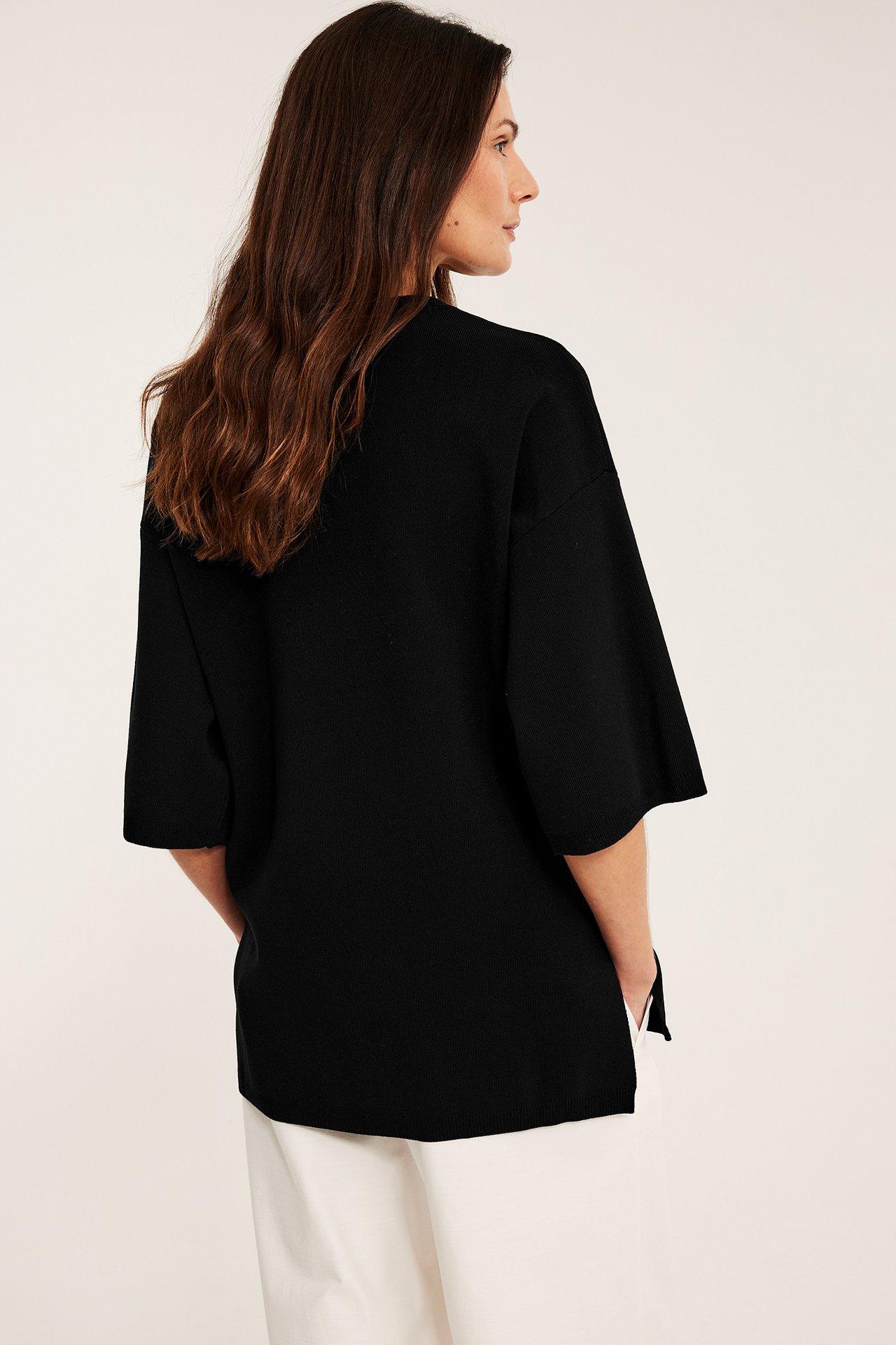 Kvinna i rygg-/sidanvy bär en svart löst sittande kortärmad finstickad tröja med ljusa vida byxor.