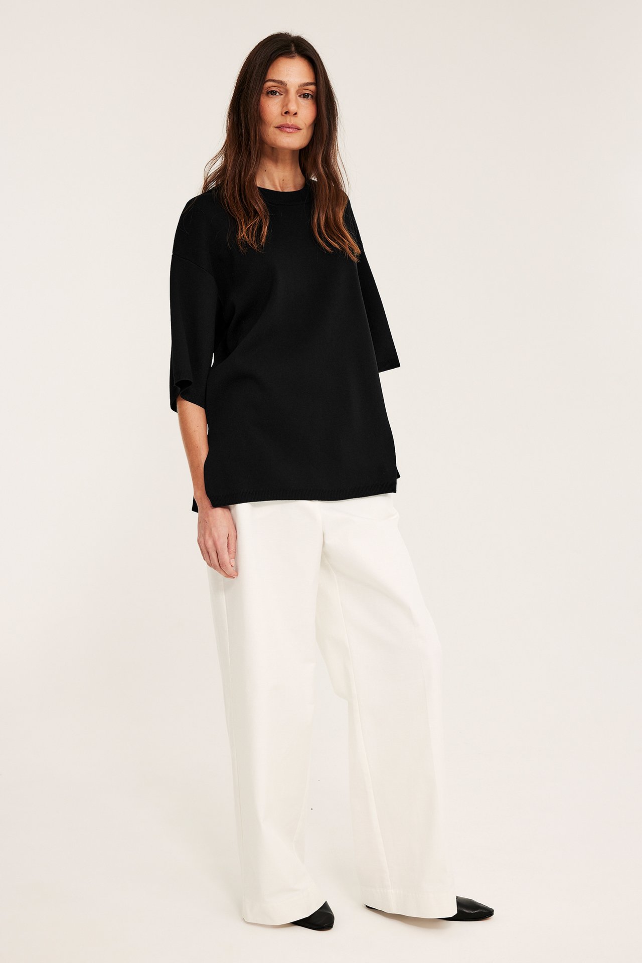 Edestäpäin: Nainen mustassa löysässä lyhythihaisessa neuleessa, valkoisissa leveissä housuissa ja mustissa kengissä.