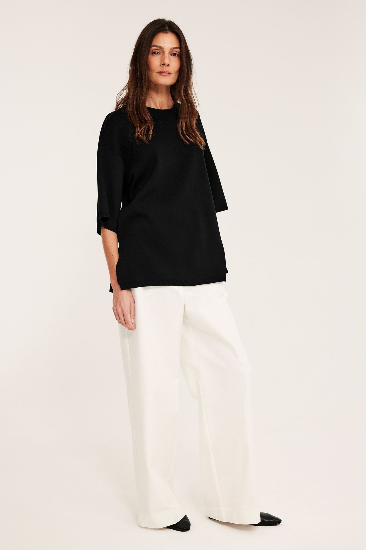 Forfra: kvinne i svart løs kortermet strikkegenser, hvite vide bukser og svarte flate sko.