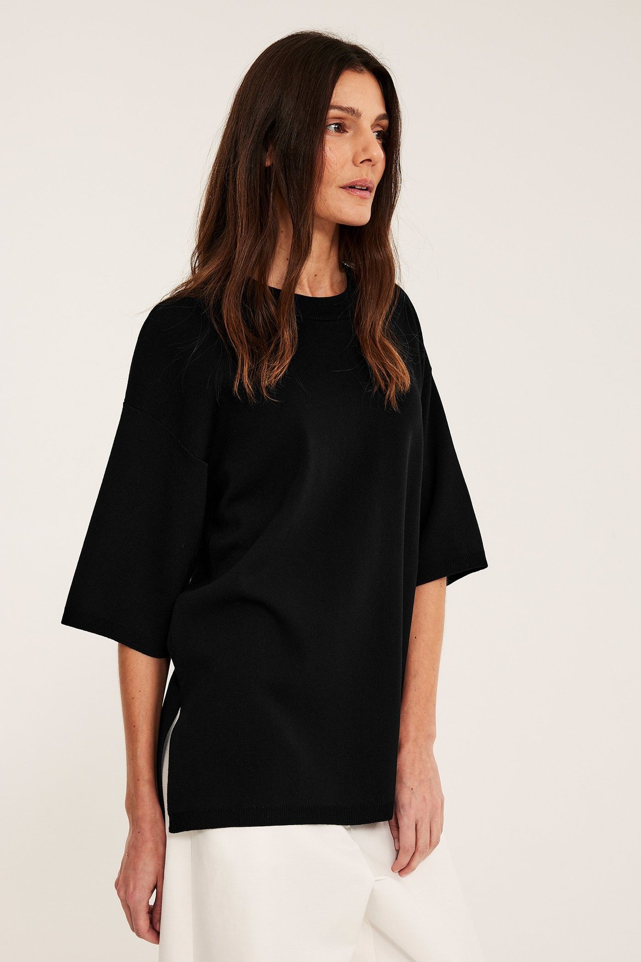 Siden av kvinne i svart løs kortarmet finstrikket genser med splitt, og hvite vide bukser.