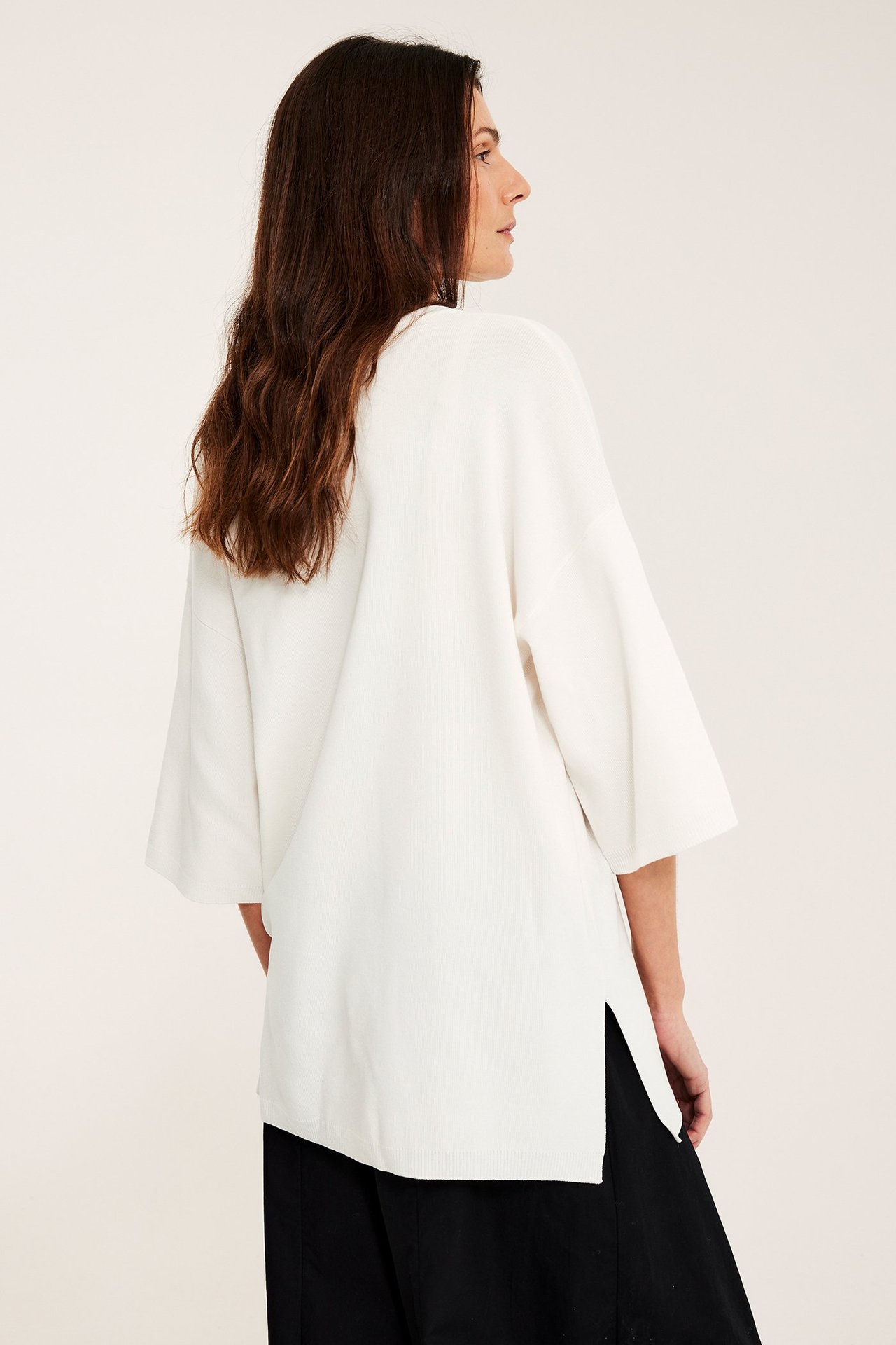 Widok z boku: kobieta w luźnym kremowym swetrze z krótkim rękawem i rozcięciami, na czarnych spodniach.
