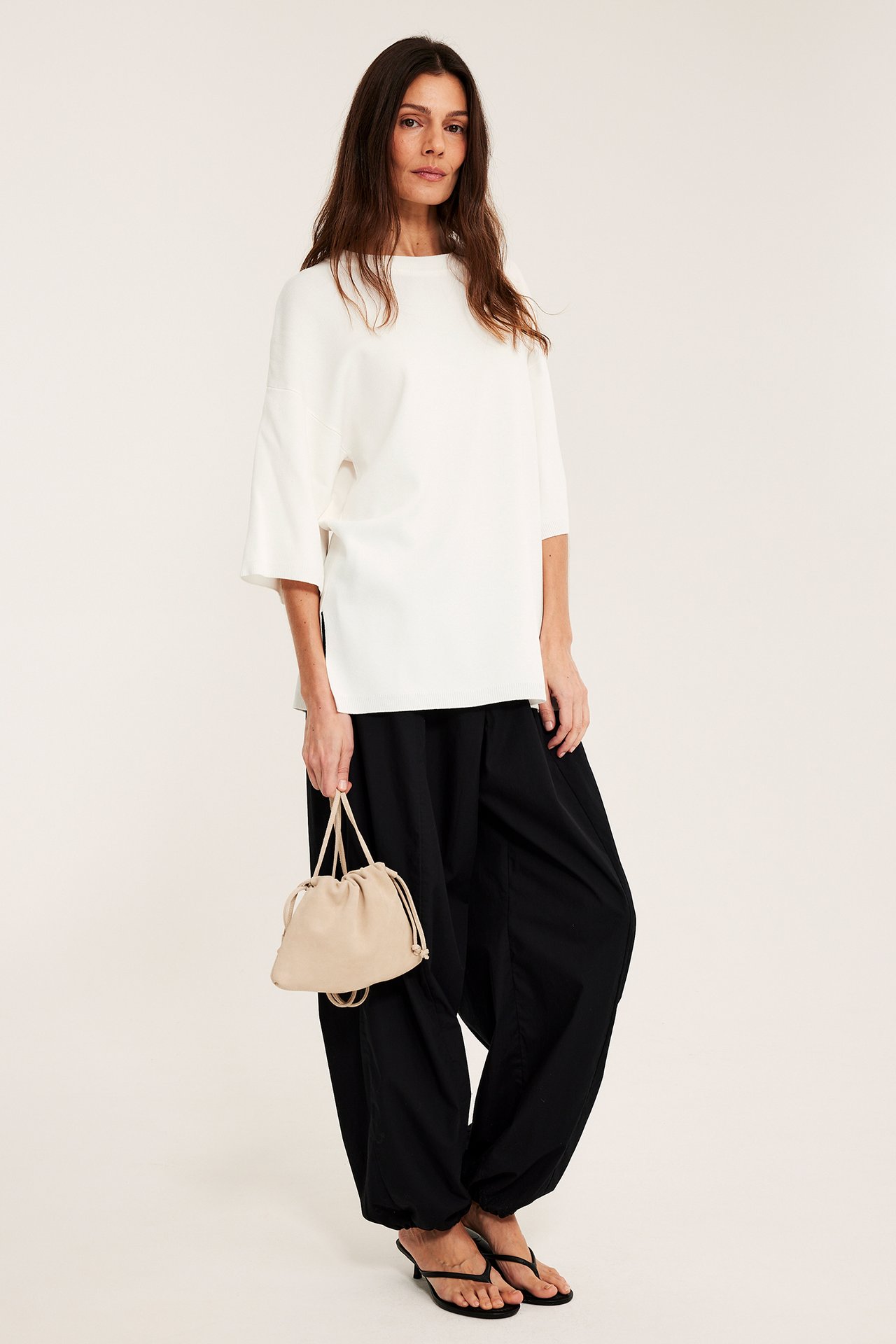 Sivukuva: Nainen luonnonvalkoisessa lyhythihaisessa neuleessa, mustat leveät housut ja beige nyörilaukku.