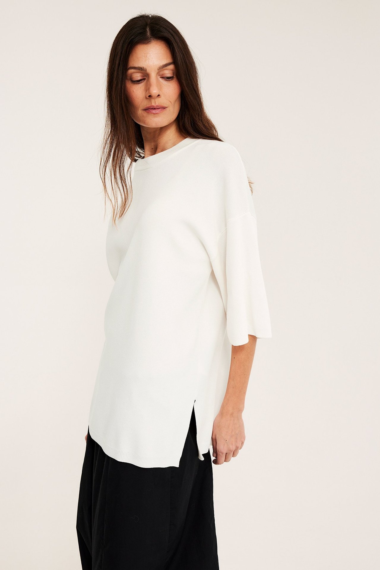 Forfra, kvinne i en offwhite finstrikket løs kortermet genser med sidesplitter over svarte løse bukser.