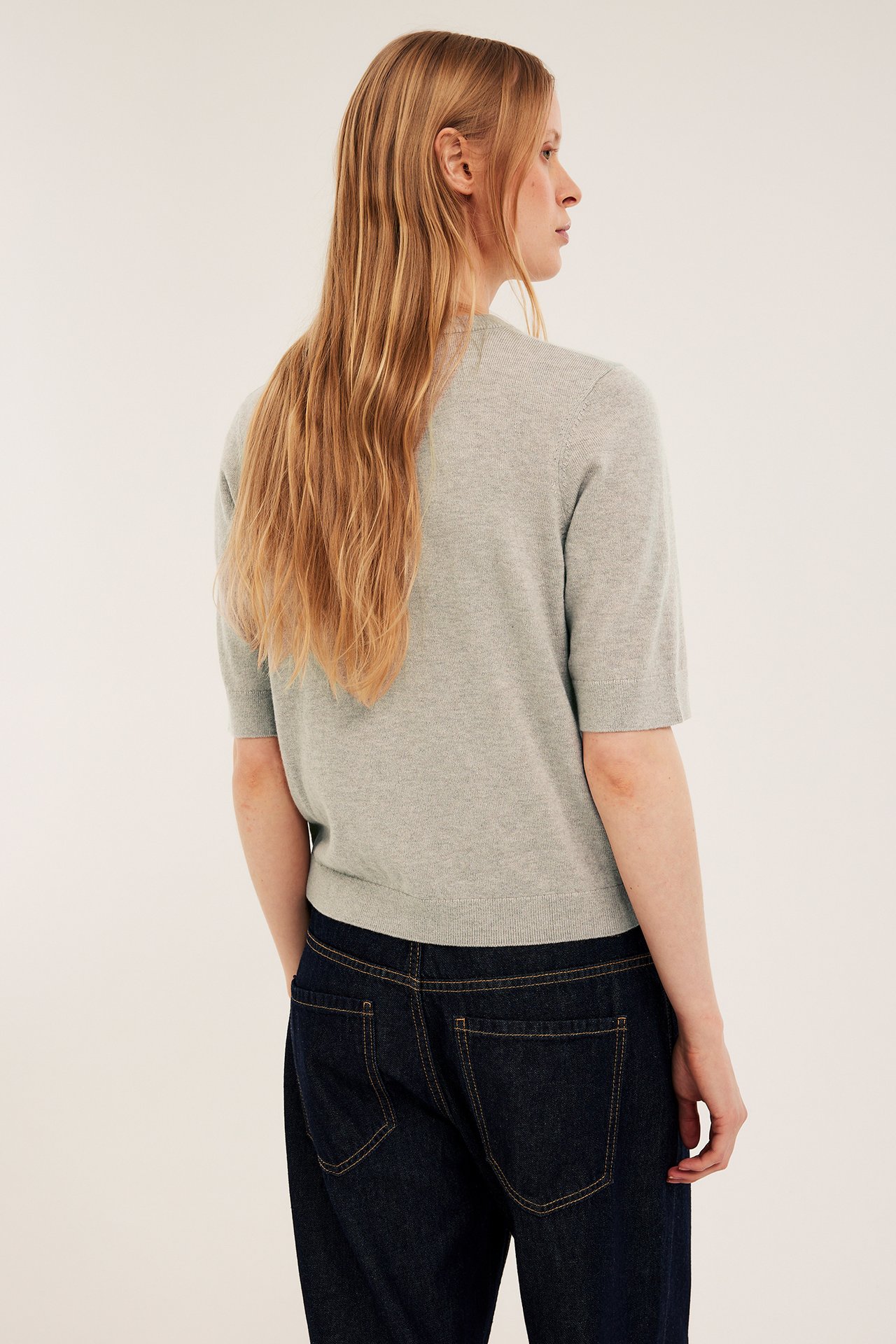 Naisen takasivukuva, yllään vaaleanharmaa meleerattu hienoneulottu lyhythihainen villasekoitepusero ja tummansiniset farkut.