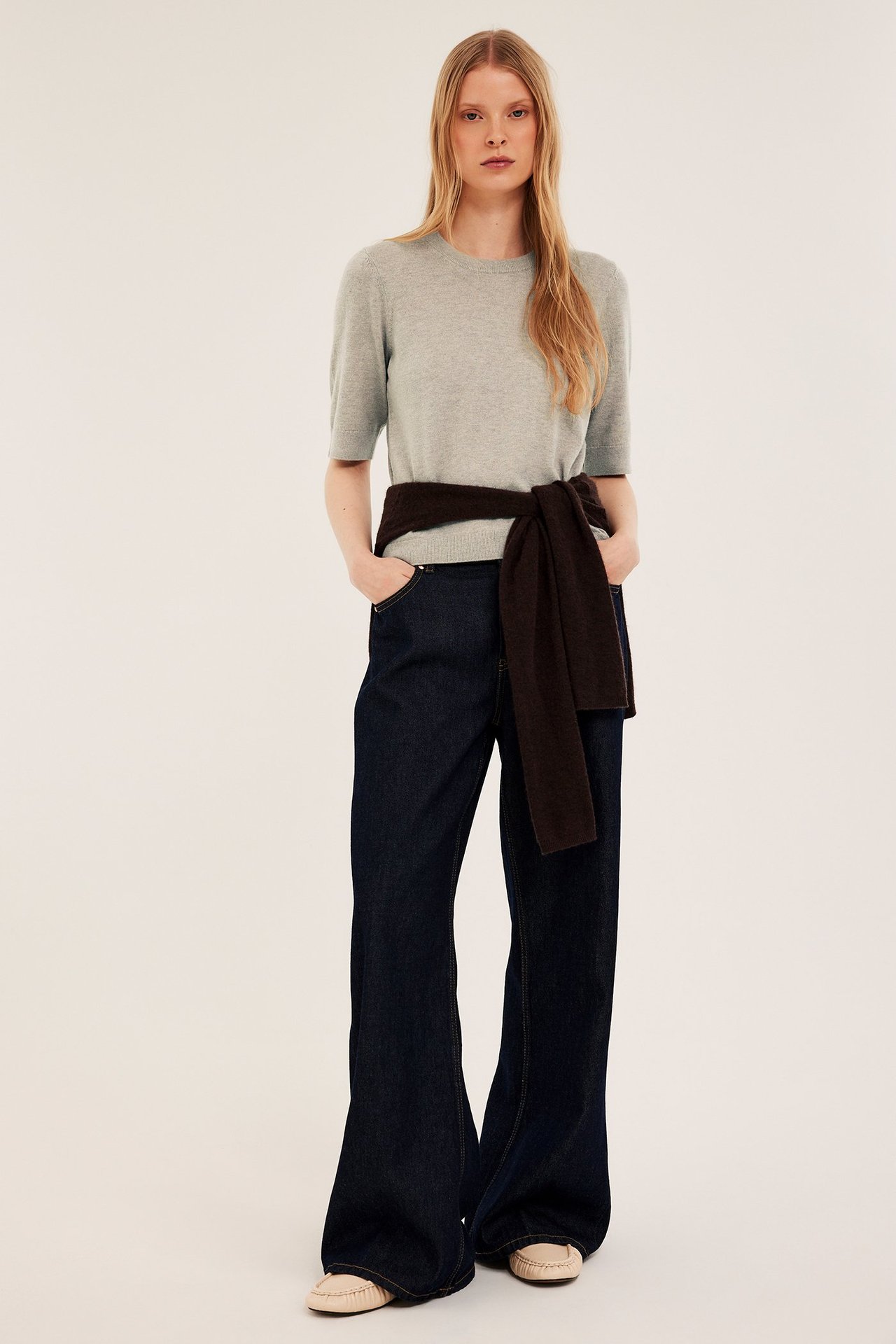 Forfra: kvinne i lys beige melert kortarmet genser, mørkeblå jeans og en brun knyttet genser.