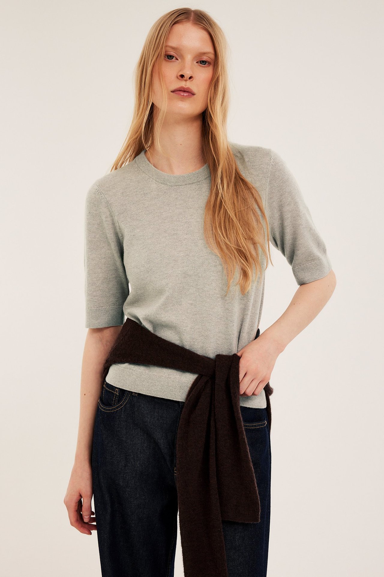 Widok z przodu: Kobieta w jasnobeżowym melanżowym swetrze z krótkim rękawem, ciemnych jeansach, brązowy sweter zawiązany w pasie.