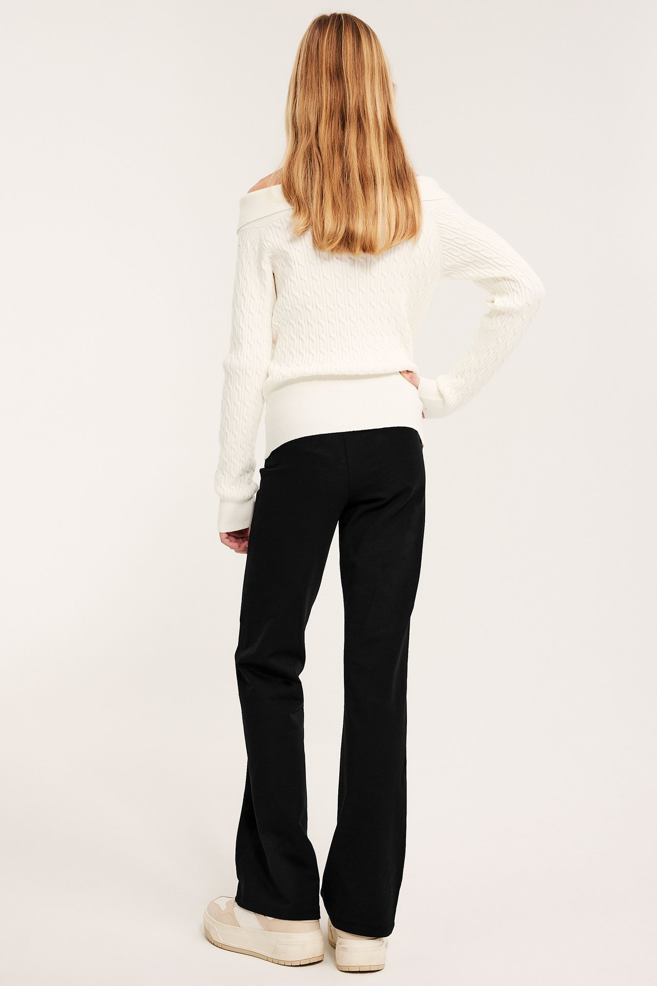 Ryggvisning av et barn i svarte flare-leggings, hvit off-shoulder kabelstrikket genser og beige sneakers.