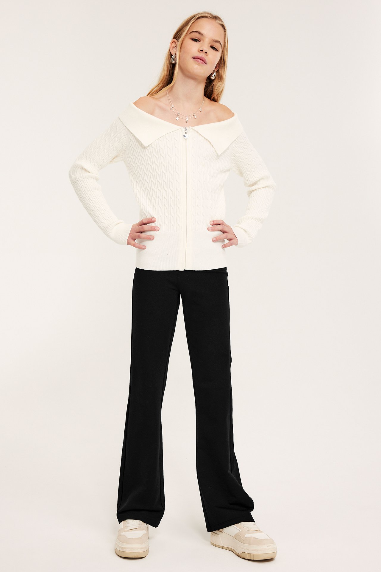 Widok z przodu dziecka w czarnych legginsach flare, kremowym kardiganie i jasnobeżowych sneakersach na platformie.