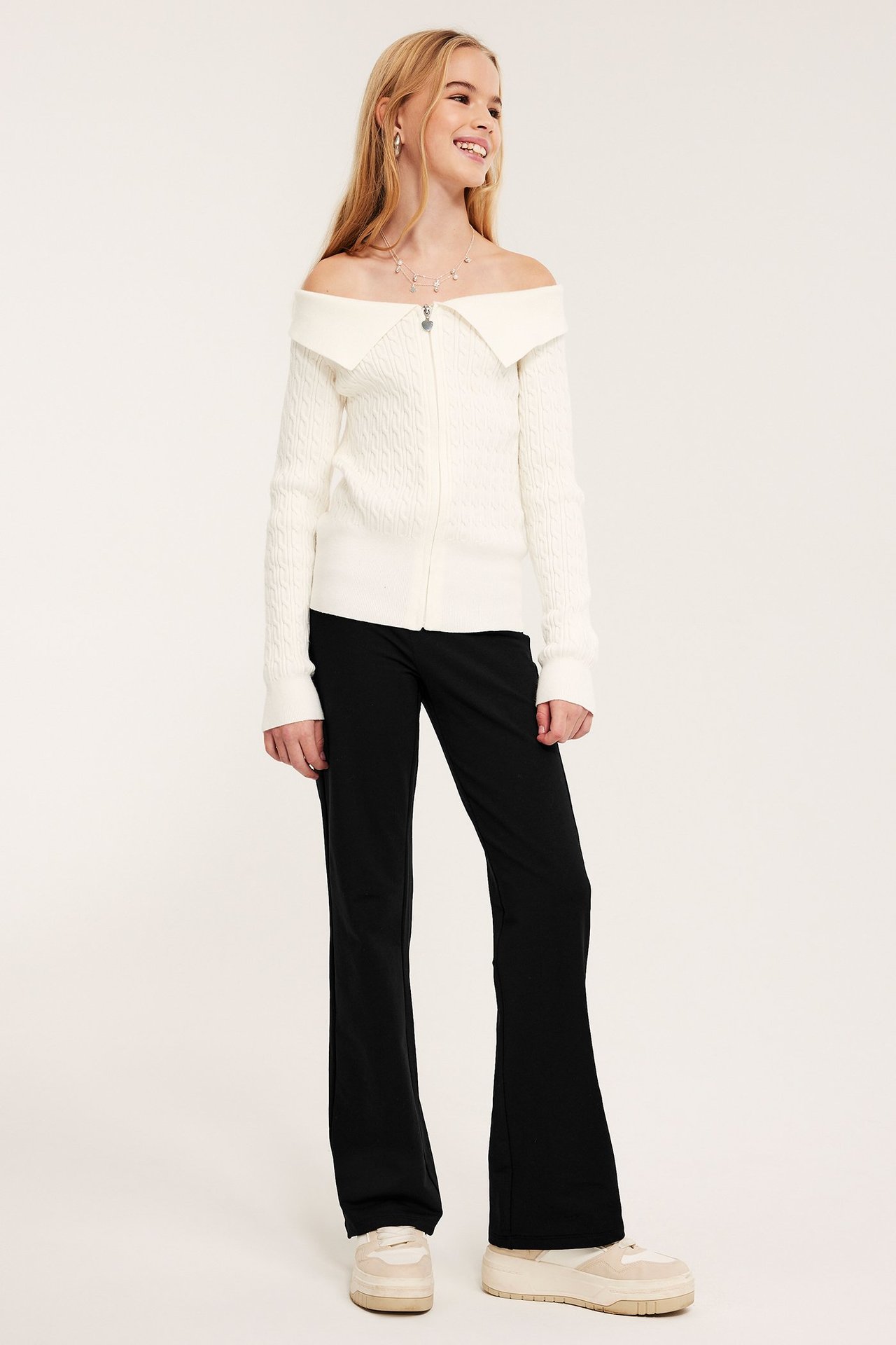 Widok z przodu: Dziecko w czarnych bawełnianych legginsach dzwonach, białym swetrze off-shoulder i beżowych trampkach na platformie.