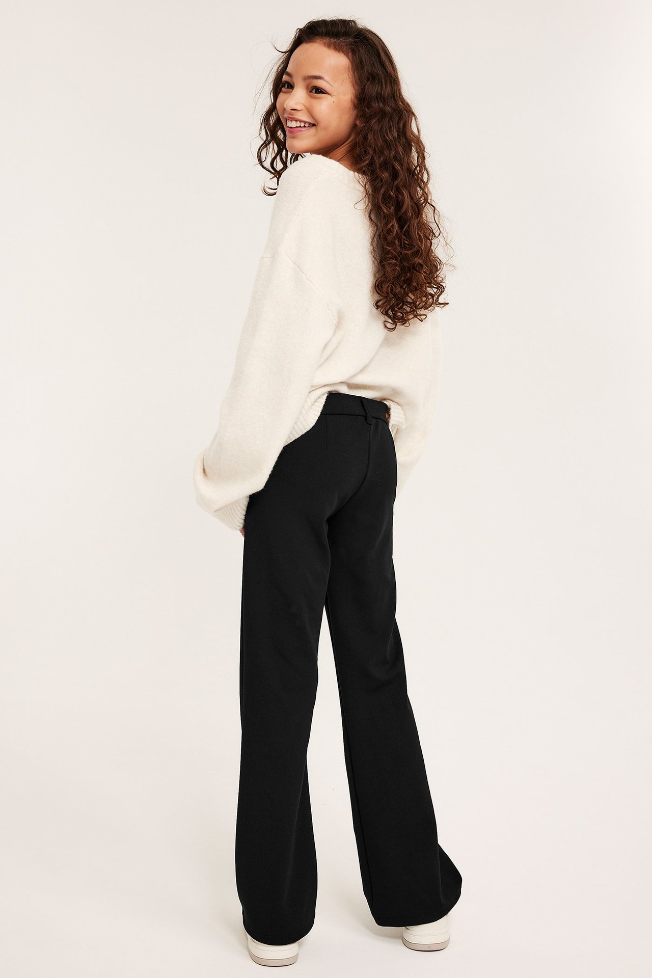 Kid i svarta utsvängda trikåbyxor och en benvit stickad tröja, sedd bakifrån.