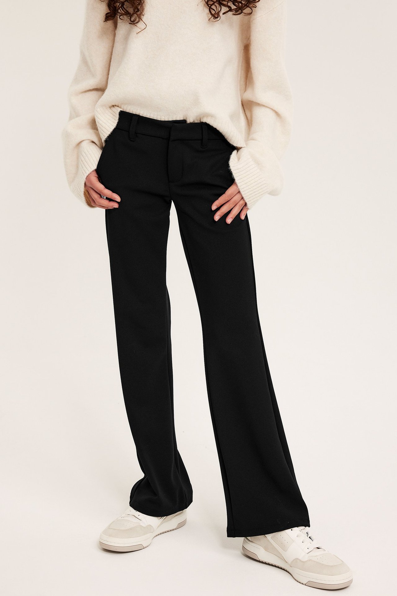 Edestäpäin, nainen mustissa levenevissä trikoohousuissa, kermanvärisessä neuleessa ja valkoisissa lenkkareissa.