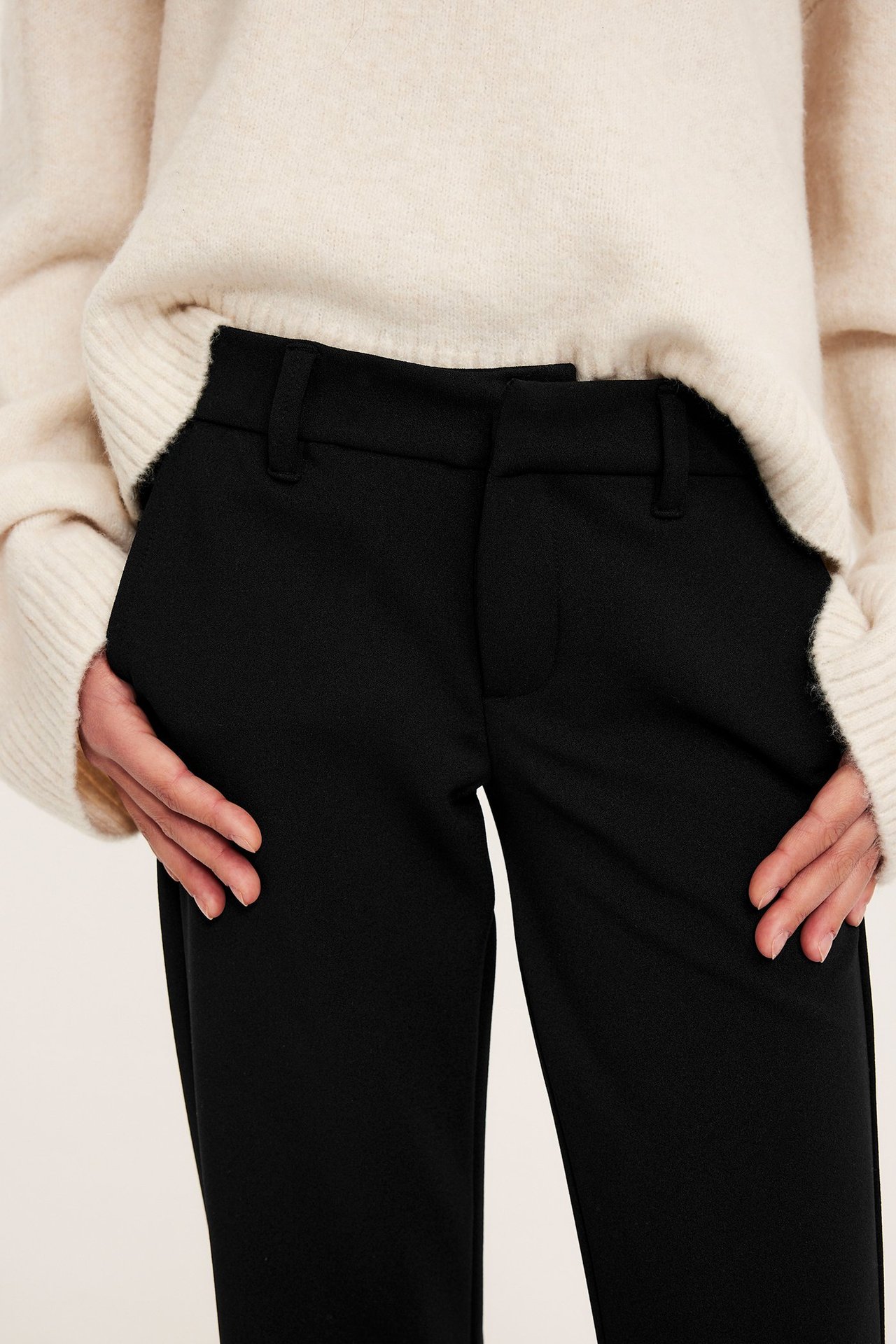 Framifrån: kvinna i svarta utsvängda trikåbyxor med händer i fickorna och en ljusbeige stickad tröja.