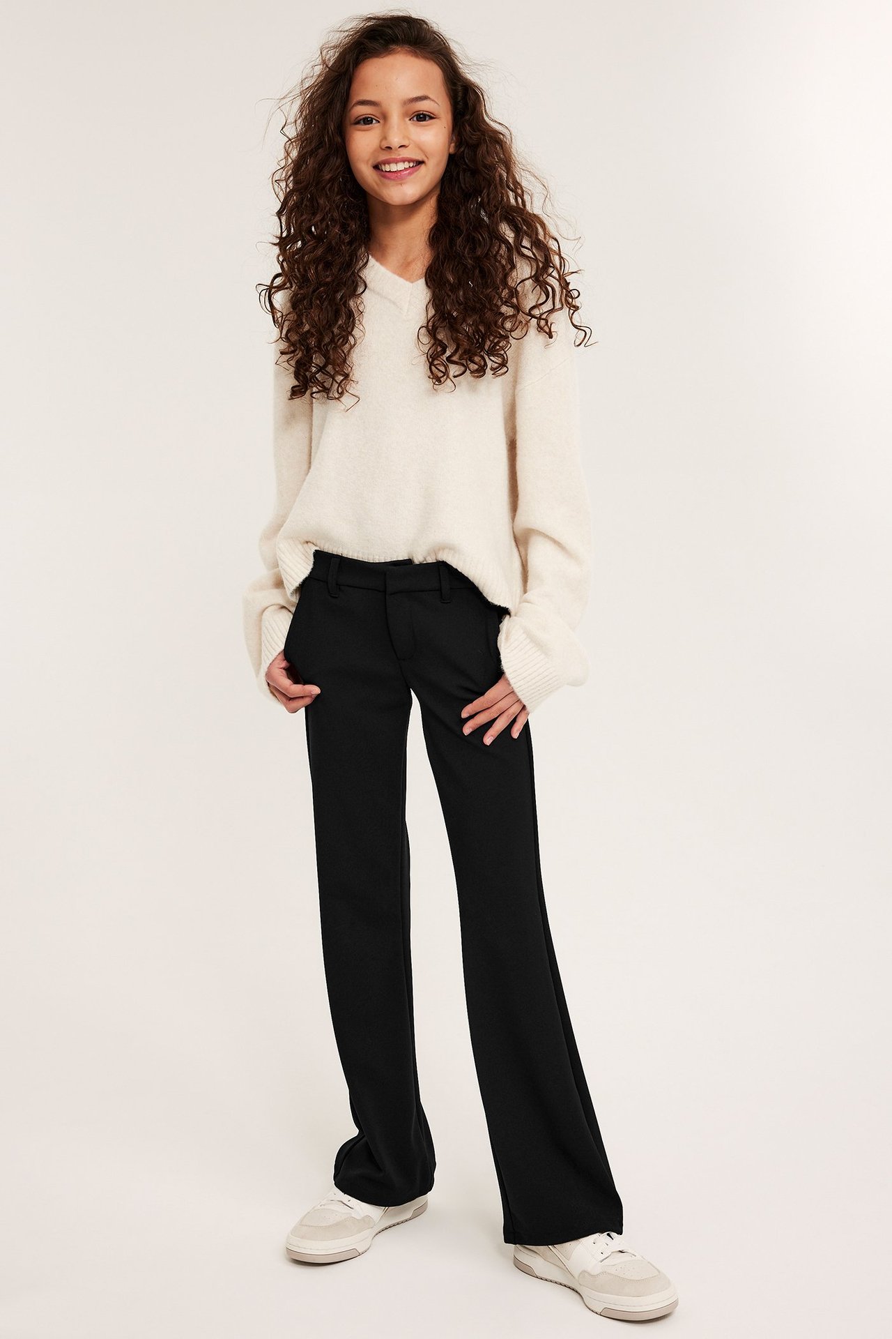 Widok z przodu dziecka w czarnych dresowych spodniach flare, beżowym swetrze i białych sneakersach.