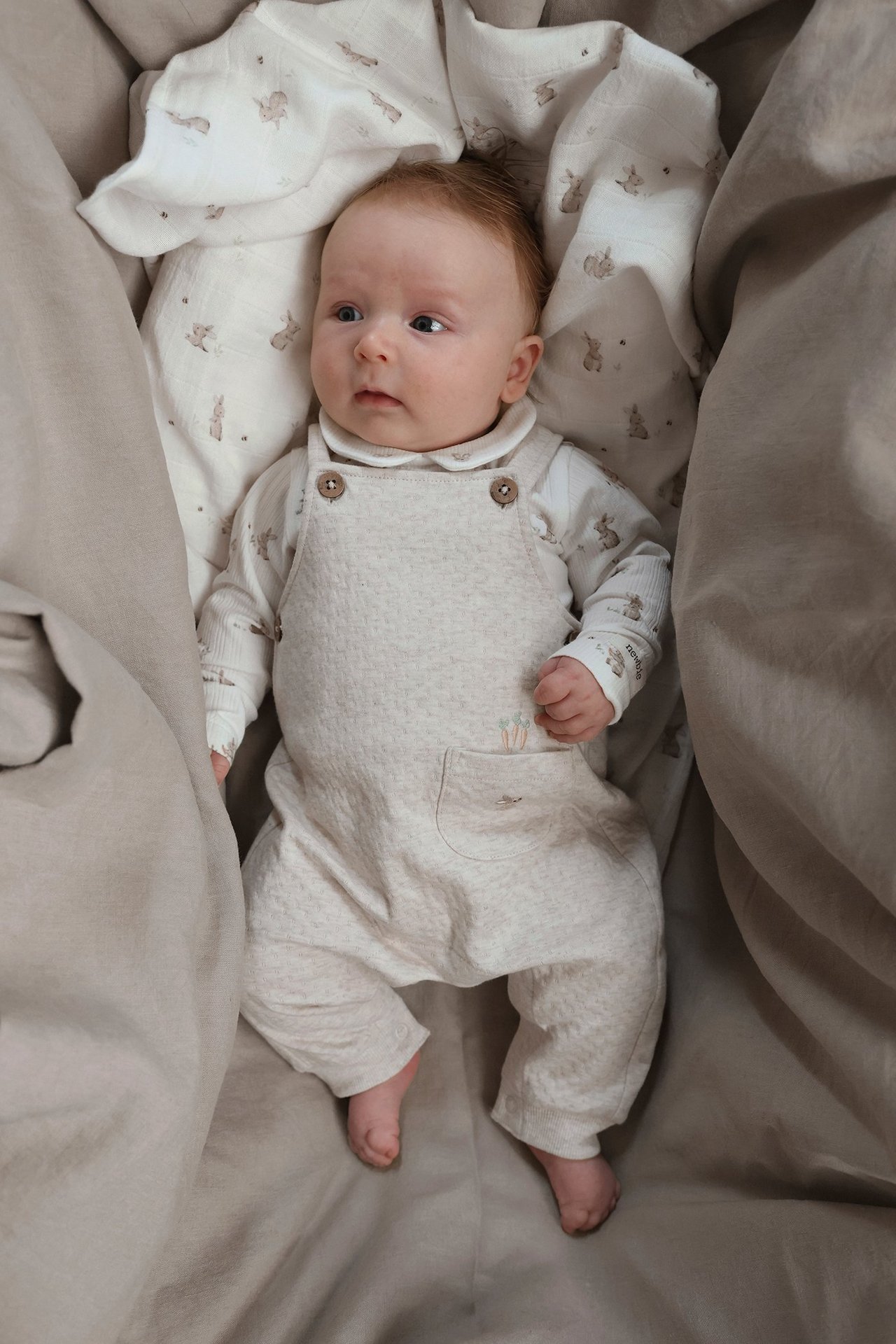 Uppifrån bild på baby i beige melerade jerseyhängslebyxor med broderad morot över vit topp med kaninmönster.