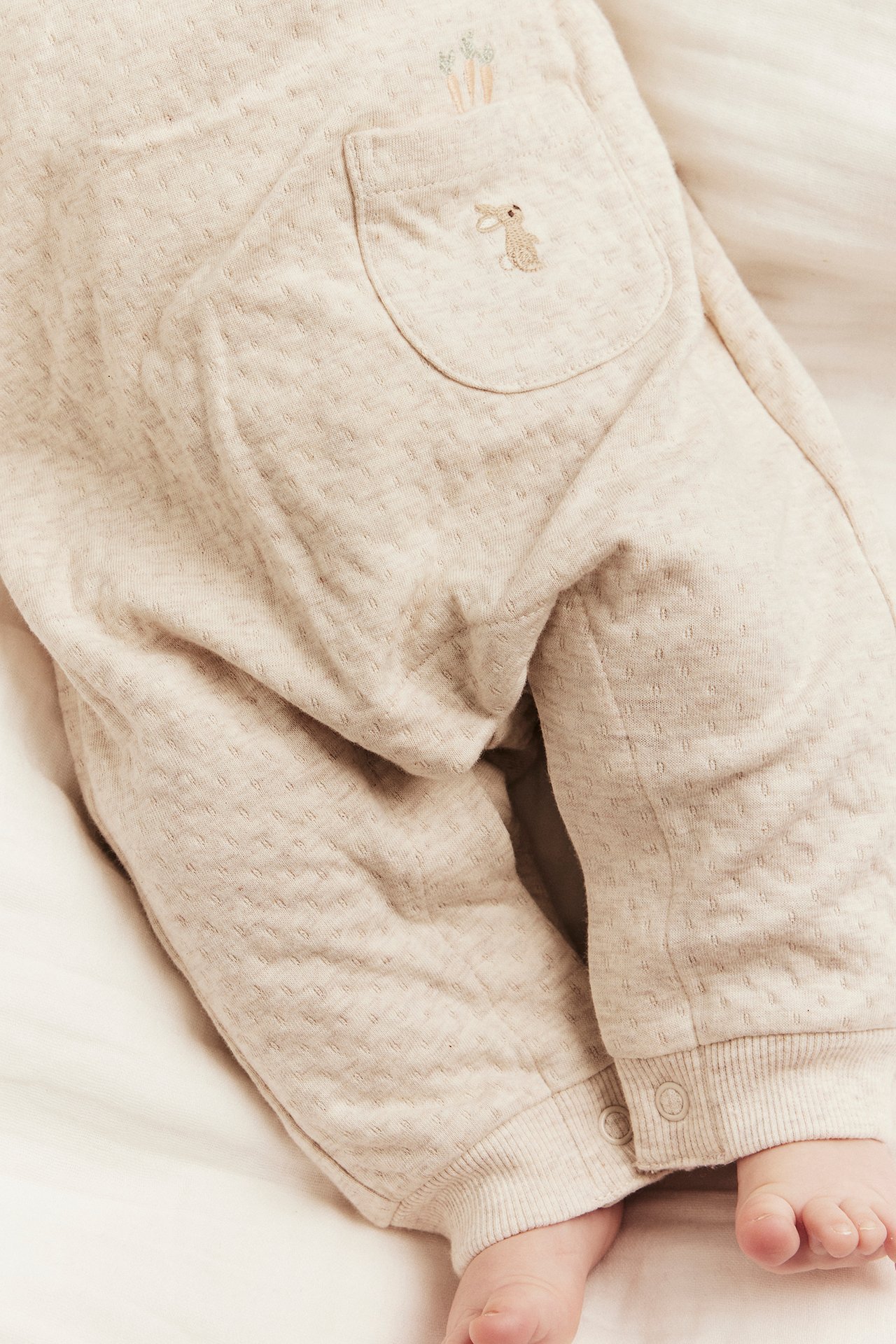 Nærbilde av baby i beige melert jersey-selebukse med brodert kaninlomme og ribbekanter.