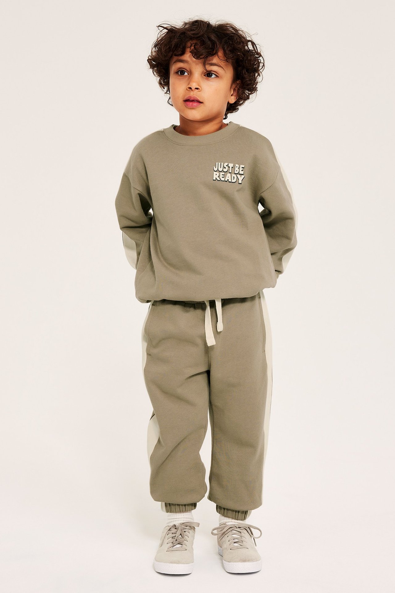 Edestäpäin: Lapsi beigen puuvillahousuissa vaaleilla sivuraidoilla, yhteensopiva paita ja beiget lenkkarit.