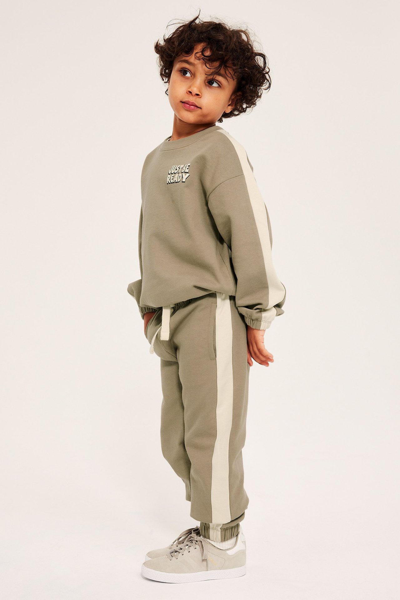 Sivukuva: Lapsi beigessä verryttelyhousuissa, joissa vaaleat sivuraidat, yhteensopiva pusero ja vaaleanharmaat lenkkarit.